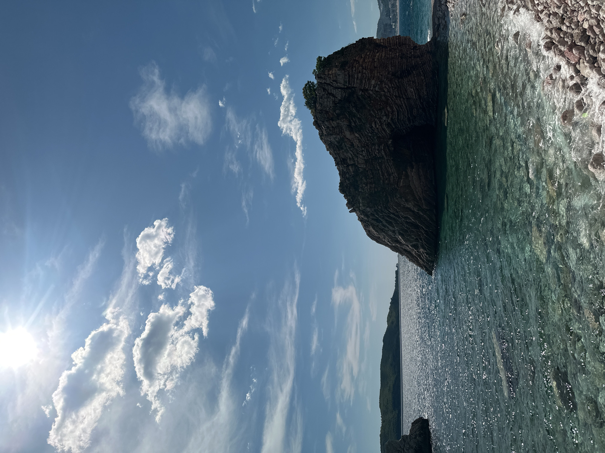 This vibrant landscape photograph captures a serene coastal scene under bright daytime sun, likely in Rafailovići, Montenegro. The image is dominated by a large, rugged rock formation or sea stack on the right, characterized by its dark, layered texture and a small cluster of green foliage crowning its peak. An arch-like opening or small cave is visible on the rock's face.
The lower and central parts of the image are filled with the clear, turquoise-green waters of the sea, sparkling brightly with sun reflections. Near the shore on the far right, gentle waves break against a pebbly or rocky coastline, revealing the underwater details through the translucent water.
In the distance, across the horizon to the left, a faint, dark silhouette of another coastline or island is visible. The sky above is a clear, light blue, dotted with various white clouds, from small wisps to larger, fluffy formations. The sun, a brilliant light source, is positioned in the upper left, casting a noticeable lens flare and bathing the entire scene in intense light, indicative of a bright, sunny day, likely mid-morning or afternoon. No people or visible text are present, emphasizing the natural beauty and tranquility of the coastal environment.
