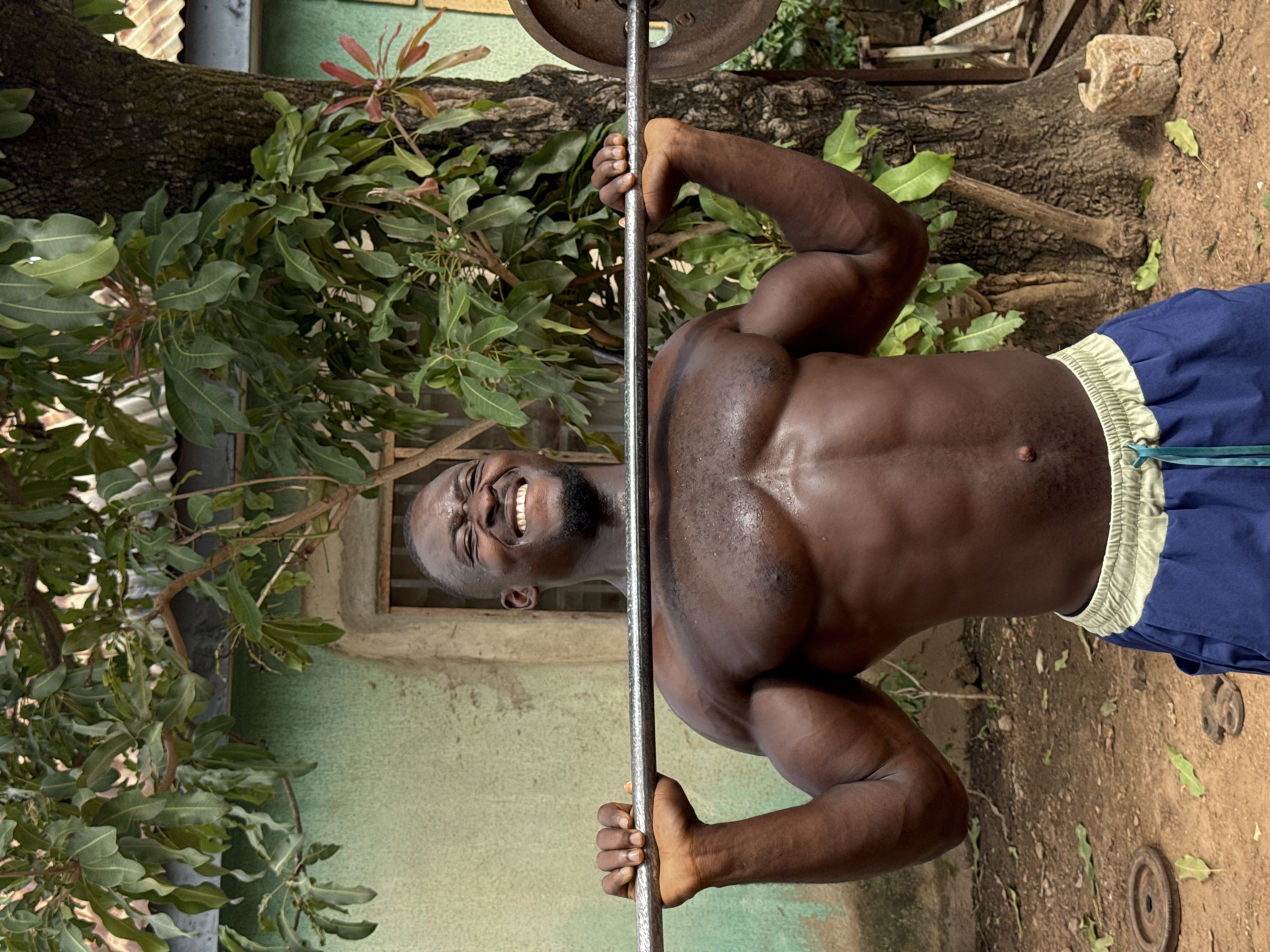 A muscular Black man, shirtless and wearing blue shorts with a light-colored waistband, is captured in a moment of intense physical exertion while weightlifting outdoors. He grips a barbell, which has a weight plate visible on one side, above his head, indicating a press or squat. His face is turned towards the viewer, featuring a wide, joyful smile that reveals his teeth, despite visible sweat on his chest and shoulders, suggesting significant effort. He has a neatly trimmed beard.
The setting appears to be a rustic outdoor area, likely a backyard or makeshift gym. Behind the man, a large tree trunk is prominent, with numerous green-leaved branches extending around him. The leaves are broad and ovate, characteristic of tropical trees. In the background, sections of a corrugated metal structure and a light green wall with a small, dark opening (possibly a window) are visible. The ground is dirt, and several loose, circular weight plates and a cylindrical stone block with a central hole (possibly an old grinding stone) are scattered near the base of the tree on the right.
The bright, natural lighting suggests it is daytime with clear weather. The man's expression conveys a mix of happiness, determination, and strength. There is no visible text in the image. The scene offers a glimpse into a dedicated fitness routine in a warm, outdoor environment.
