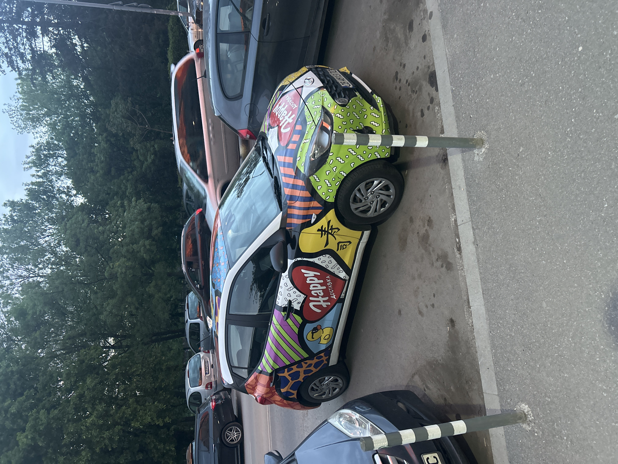 The image depicts a parking scene in Sofia, Bulgaria. Several cars are parked alongside a road with a line of trees in the background. The focus is on a brightly decorated car with a white base and various colorful patterns and designs, including the word "Happy" in Cyrillic. The car is parked very close to a metal bollard with black and white stripes. Other cars, including a gray hatchback, a silver sedan, and a dark-colored sedan, are visible in the background and foreground. The road appears to be asphalt, and the lighting suggests an overcast or late afternoon.