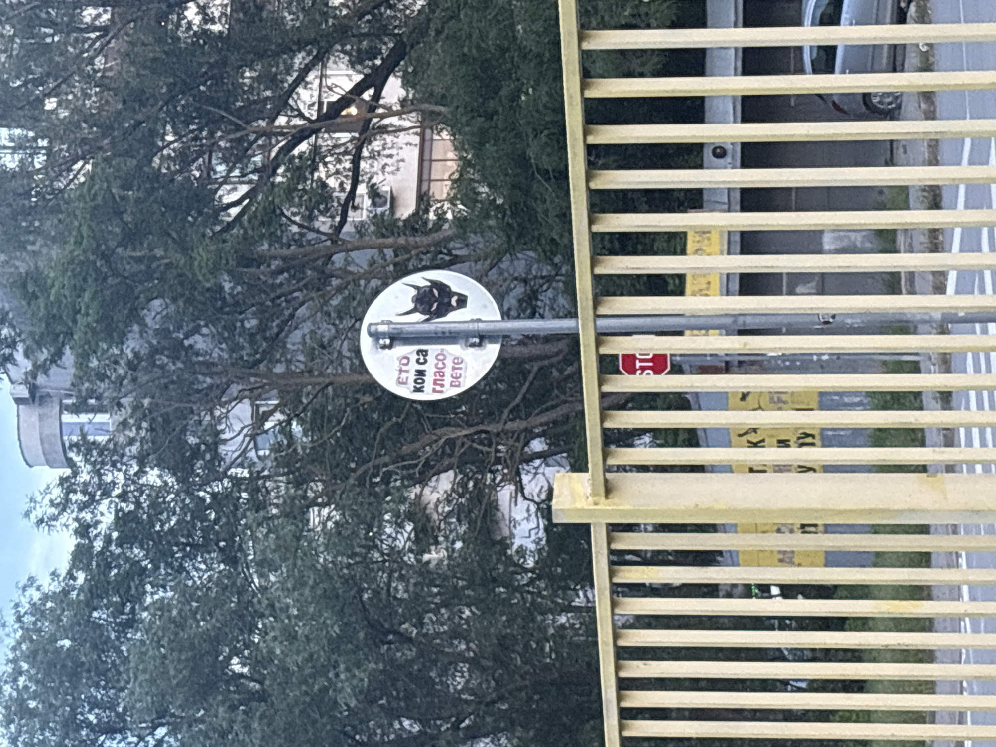 A daytime outdoor scene captures a street view from behind a yellow metal fence. The fence, composed of evenly spaced vertical bars and a horizontal top rail, occupies the lower third of the image. Behind the fence, a white circular sign is mounted on a metal pole. The sign displays Cyrillic text in black and red on its left side, reading "ЕТО КОИ СА ГЛАСОВЕТЕ," and a black graphic of a stylized horned head or demon mask on its right. Below this, a red octagonal "STOP" sign is partially visible. Further back, a road or paved area is present, with faint white markings on the ground. A silver car is partially visible on the right, parked or driving. In the midground, beyond the fence and signs, there are additional structures; one appears to have a yellow sign with partially visible Cyrillic text through the fence bars. The background is dominated by dense green trees with visible branches and foliage. Through and above the trees, several buildings are visible, including one with a distinct curved, light-colored upper section and rectangular windows, and another lighter-colored building showing an air conditioning unit. The sky above is light blue with some cloud cover. No people are visible in the image.