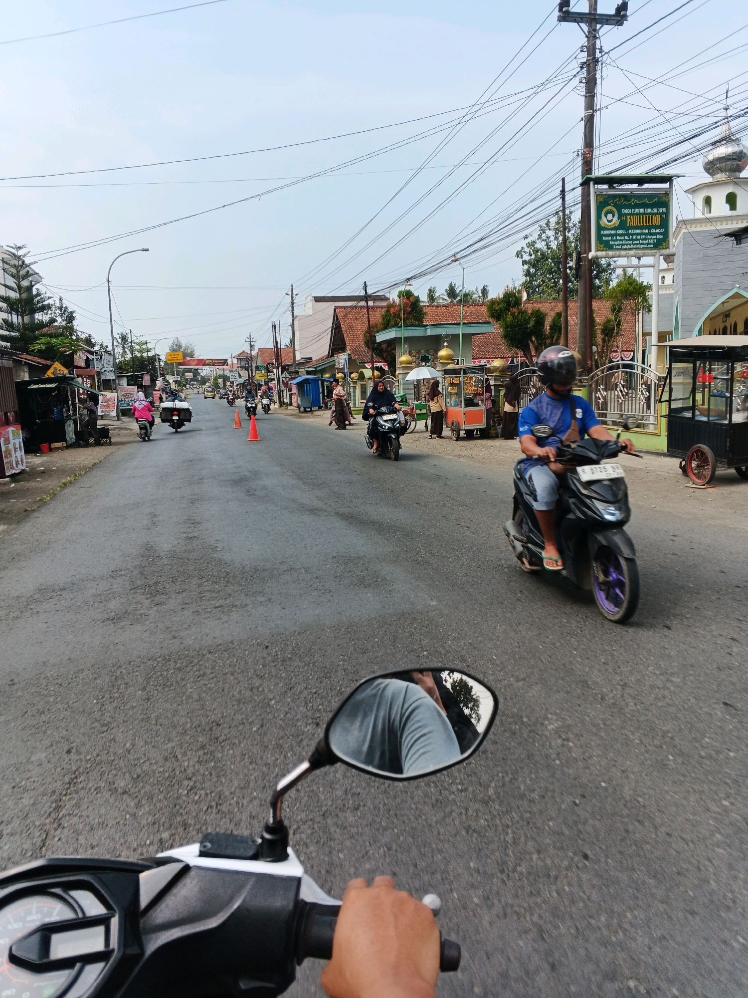 The image depicts a street scene in Jangrana, Indonesia. A person is riding a black scooter on the right side of a paved road. The rider is wearing a blue shirt, a dark helmet, and light-colored shorts. A scooter mirror reflects the rider and a partial view of the sky. Several other motorcycles and scooters are visible on the road, along with some pedestrians. Buildings line the street, including a mosque with a dome and minaret on the right side of the image. Street vendors with carts are also present. The sky is overcast, and power lines crisscross above the street.