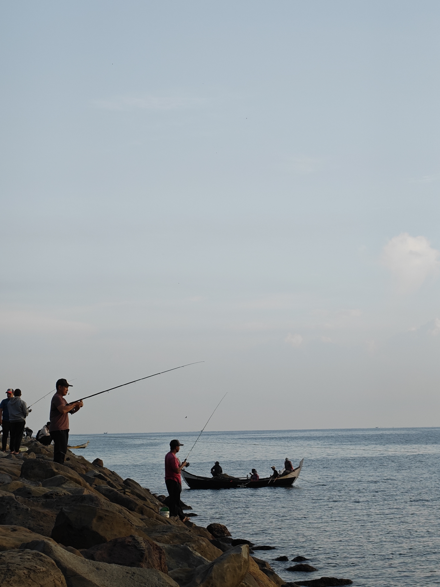 This image captures a scene in Lhokseumawe, Indonesia, where several individuals are engaged in fishing activities. Some people are fishing from a rocky shoreline, with fishing rods extended over the water. A small boat is also present in the water, occupied by a group of people. The sky is light with a slight haze, indicating either early morning or late afternoon light. The sea appears calm. The rocky shoreline is composed of large boulders.