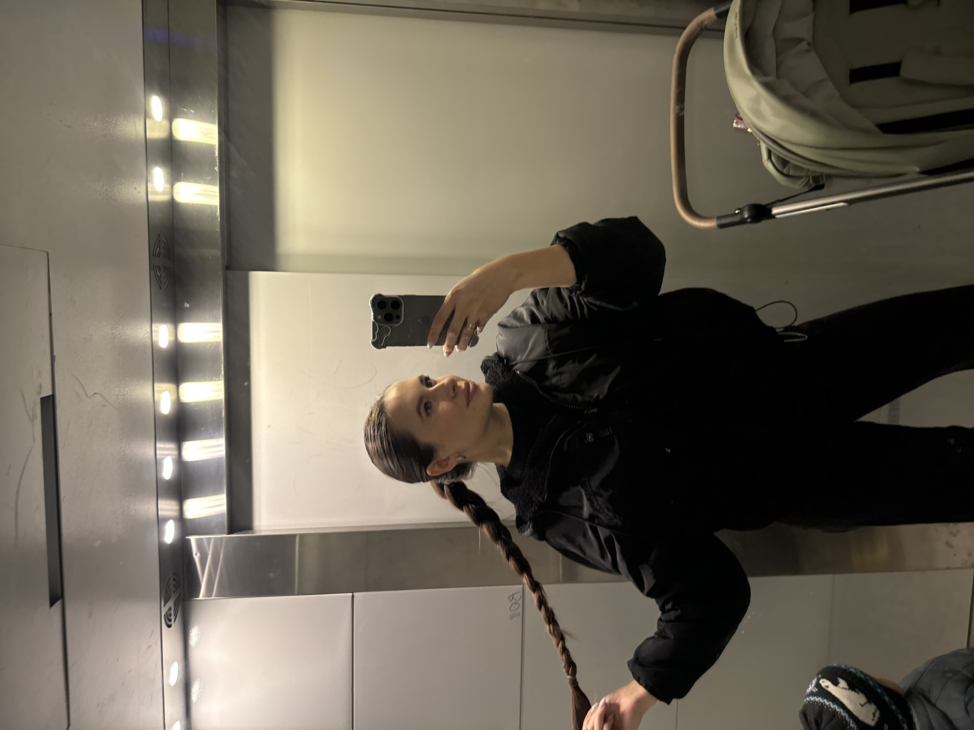 A woman in a black jacket and black pants is taking a selfie in a mirrored elevator. She is holding a smartphone in her right hand. Her hair is styled in a long braid. A stroller is visible behind her. The elevator has a panel with vertical lights. The location is likely a building in Odintsovo, Russia.