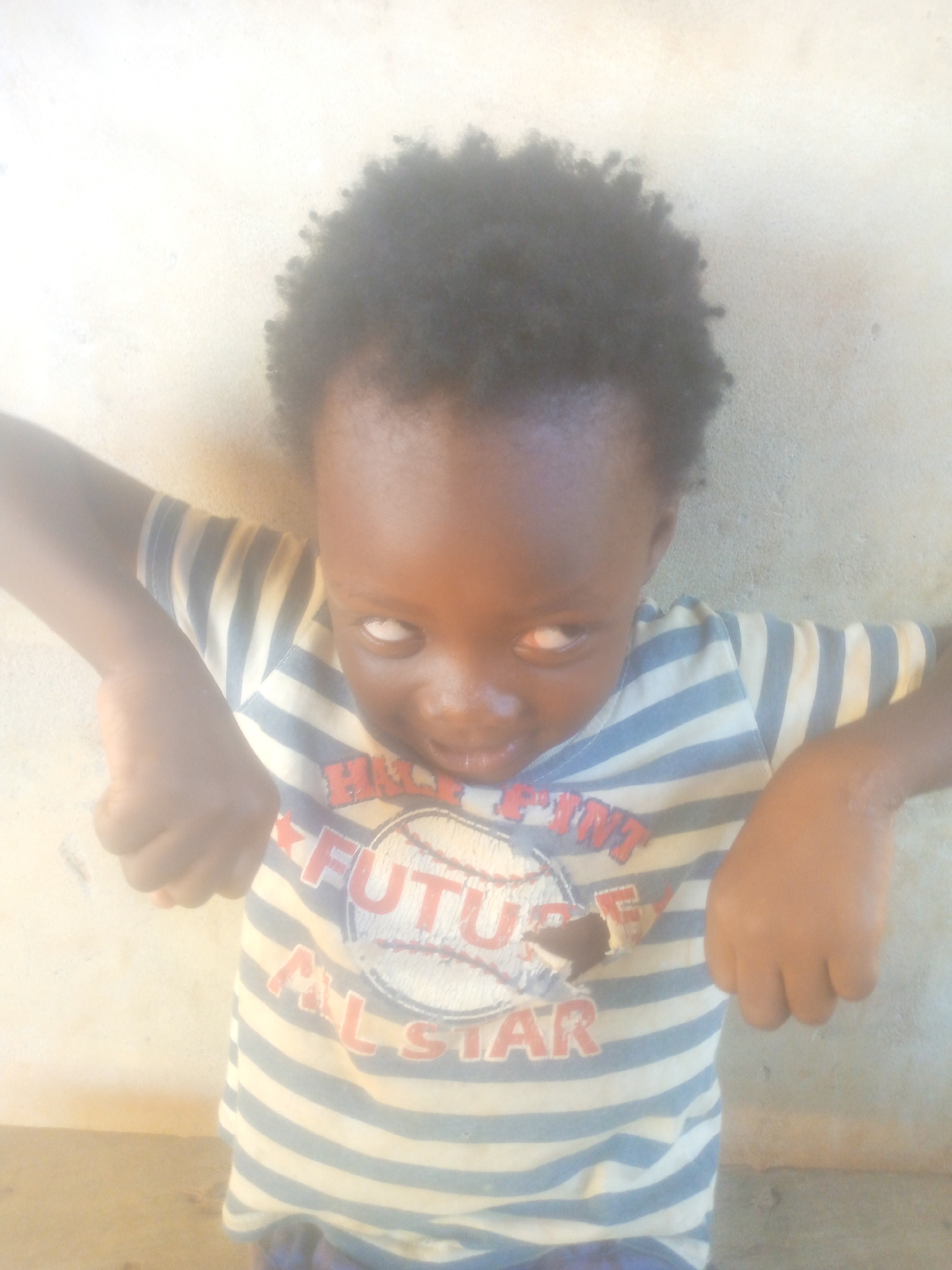 A young child with dark skin and short, curly black hair is positioned in front of a neutral-colored wall. The child wears a striped t-shirt with a white baseball design and red lettering that says "Future All-Star." The child's arms are bent at the elbows, with clenched fists held up to the sides of the head. The location is Unknown, Unknown.