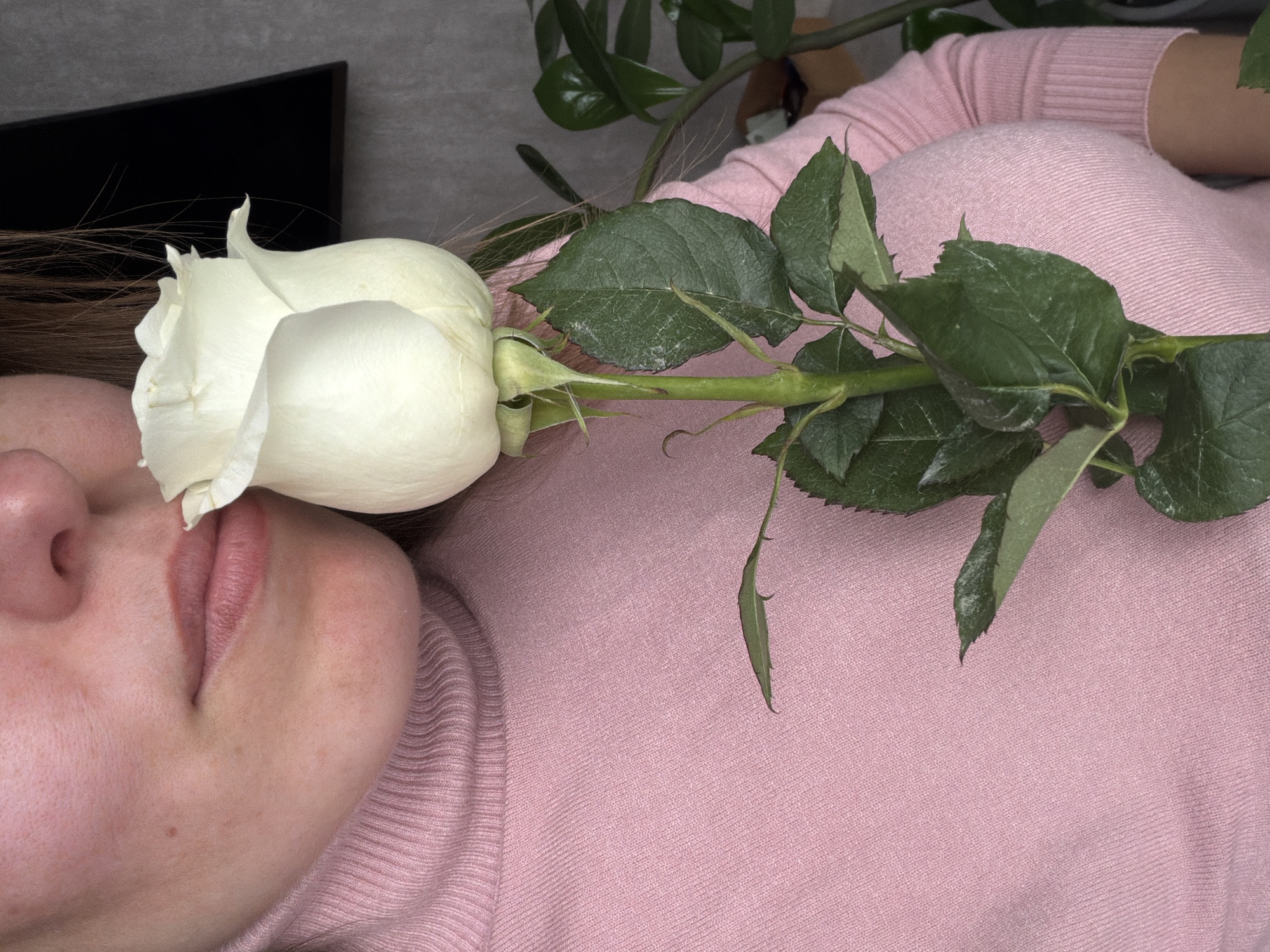 A woman in a pink sweater is lying on a gray surface. A white rose lies on her face, covering her eye. A black television is visible in the upper left corner of the image. Green leaves from a plant are visible in the upper right corner. It is not possible to determine if the location is Izhevsk, Russia.