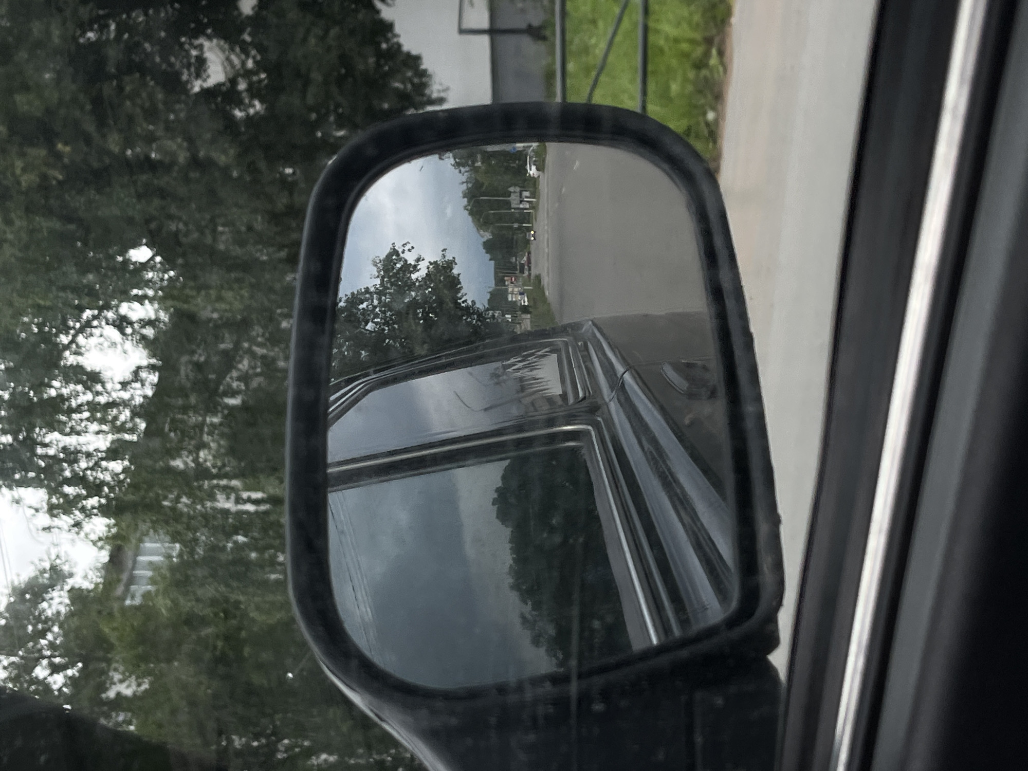 The image shows a view through the side mirror of a car in Chaikovsky, Russia. The mirror reflects a road with vehicles, trees, and buildings. The sky is overcast, indicating a cloudy day. The car appears to be in motion, as evidenced by the blurred background. The overall scene suggests a typical urban or suburban setting.