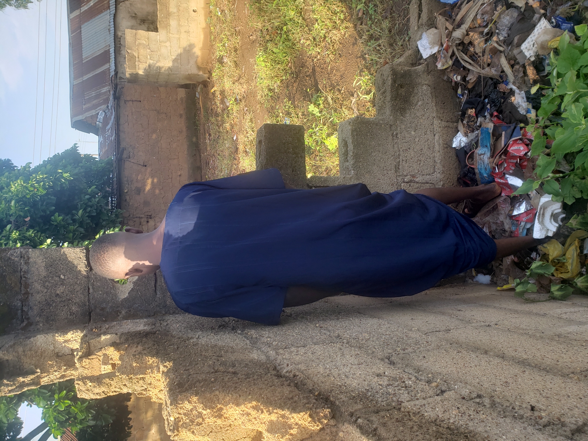 In Kachia, Nigeria, a person stands with their back to the camera, partially concealed by a weathered, crumbling concrete wall. They are wearing a dark blue, ankle-length garment. To the right of the person, a pile of refuse is visible, consisting of plastic bags, food wrappers, and other assorted debris. In the background, more dilapidated structures with corrugated tin roofs and mud brick walls are present, along with overgrown vegetation and dry grass. The sky above is clear, suggesting it is daytime.