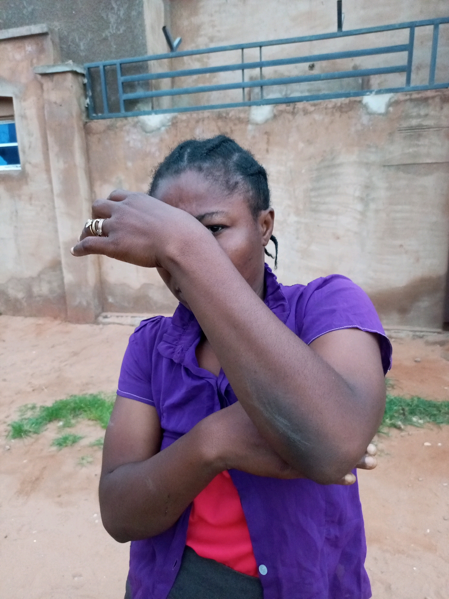 A medium shot captures a Black woman from the chest up, standing outdoors in what appears to be a rural or less developed urban setting. She is wearing a vibrant purple, collared shirt over a red undershirt. Her hair is styled in braids pulled back from her face. Her right arm is raised, covering the upper half of her face, with her hand resting on her forehead. Two rings are visible on her left index finger. The background consists of a textured, light brown wall, possibly part of a building, with a blue-barred window to the left. A gravel or dirt ground with patches of green grass is visible in the foreground. The lighting suggests it is daytime. The scene is consistent with an outdoor environment in Tempure, Nigeria.
