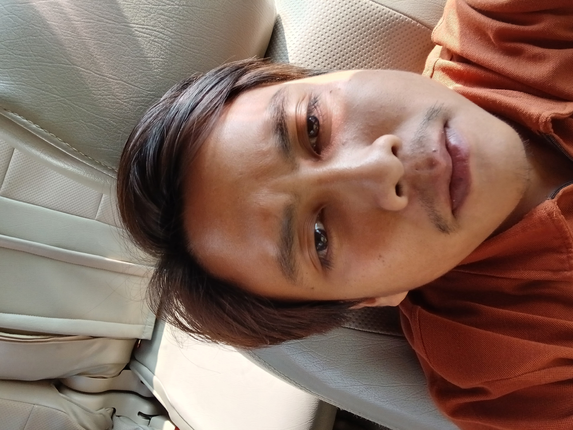 A close-up, selfie-style image shows the face of a man with brown hair and medium-toned skin, wearing an orange collared shirt. The man is seated in a car, with the beige leather interior of a car seat visible behind him. The lighting suggests it is daytime, with sunlight casting shadows on the man's face. The man's features include dark eyes, eyebrows, and a faint mustache. The background shows the textured pattern of the car's upholstery. There are no other individuals or objects of note in the frame. The image is taken from a slightly upward angle. The location is stated to be in Gombong, Indonesia.