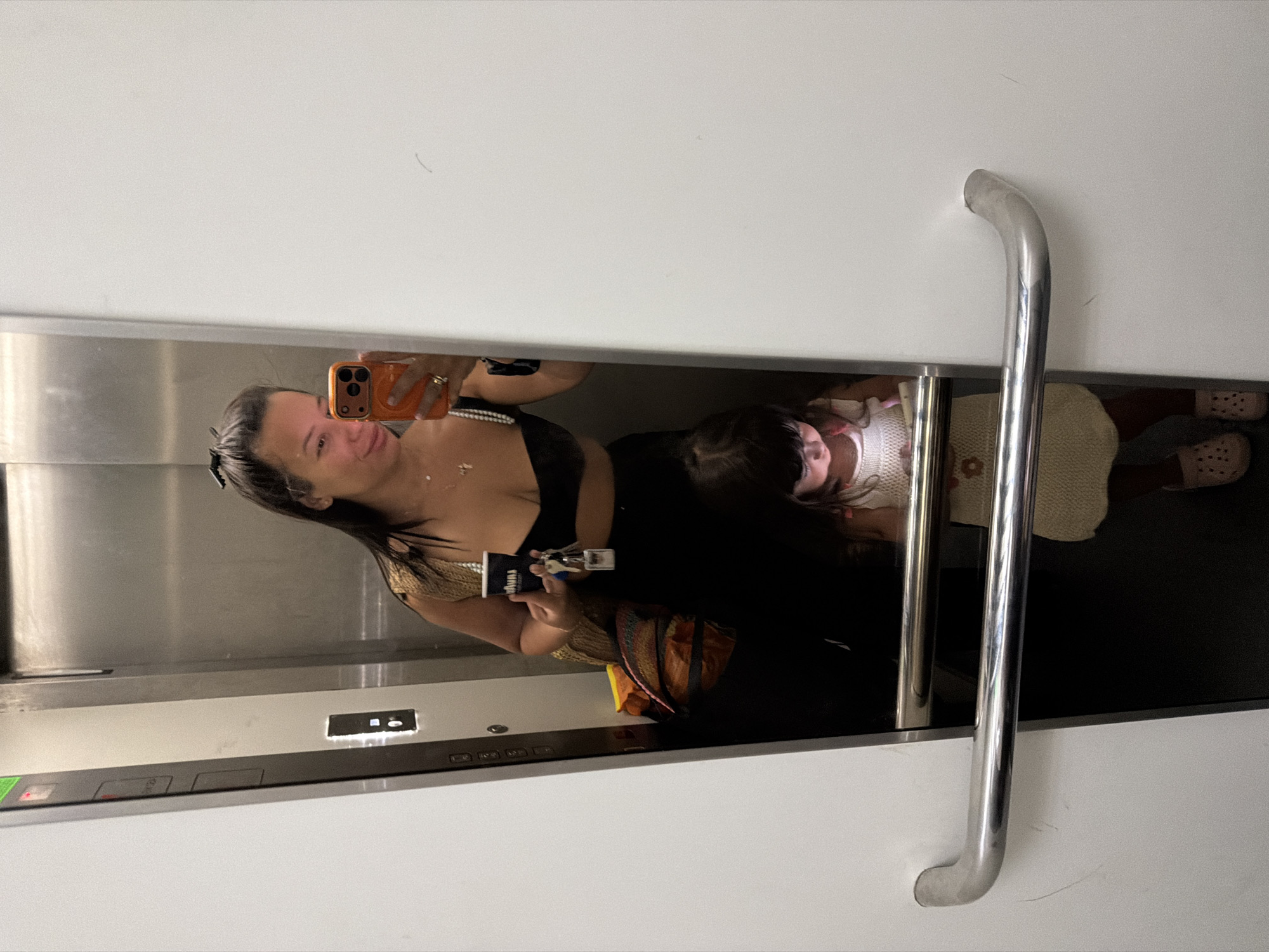 In an elevator, a woman and a young child are reflected in the mirrored walls. The woman, holding a coffee cup and keys, is taking a selfie with her phone. She is wearing a black top and jewelry. The child, dressed in a cream-colored outfit, is looking down at a lit-up device. The elevator appears to be in a modern building, possibly a hotel or residential complex, with stainless steel accents and a white interior. The location of Burgas, Bulgaria, is not discernible from the image itself.