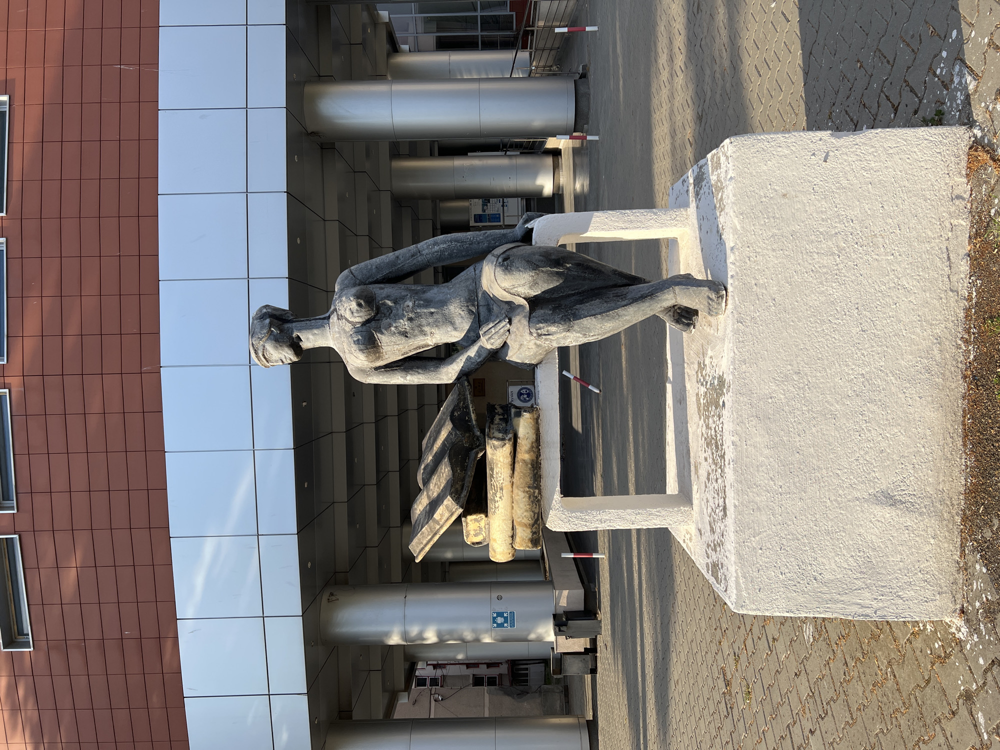 The image shows a stone statue of a woman sitting on a bench with books in front of a modern building with a red tiled facade and silver panels. The statue is located in front of the "Ovidius" University of Constanța, Faculty of Medicine. The woman is depicted with her legs crossed, looking upwards. Behind her, there is a stack of large stone books on the bench. The ground is paved with grey interlocking bricks. The building has large silver pillars supporting an overhang. The lighting suggests it is daytime, with shadows cast on the ground. There are trash bins to the left of the statue and two bollards with red and white markings in front of the bench. A sign with a blue and white emblem is visible on one of the pillars.