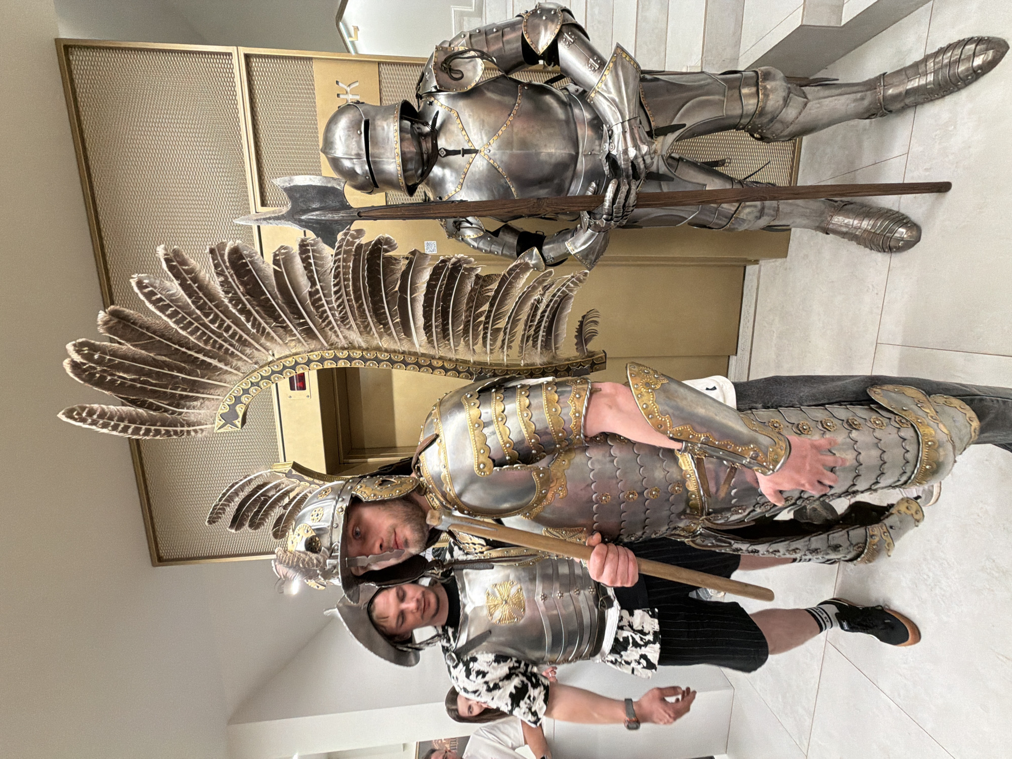 The image features three people and a suit of armor in an indoor setting.
Two men stand in the foreground. One wears a partial suit of winged Hussar armor with a black and white patterned shirt and black and white striped shorts. The other man wears a hat and a vest that is part of a suit of armor over a black shirt. Both men hold wooden polearms.
In the background, a full suit of armor lies on the floor. It also holds a polearm. A textured gold panel is behind the armor.
The location and city are not clear from the provided image.
