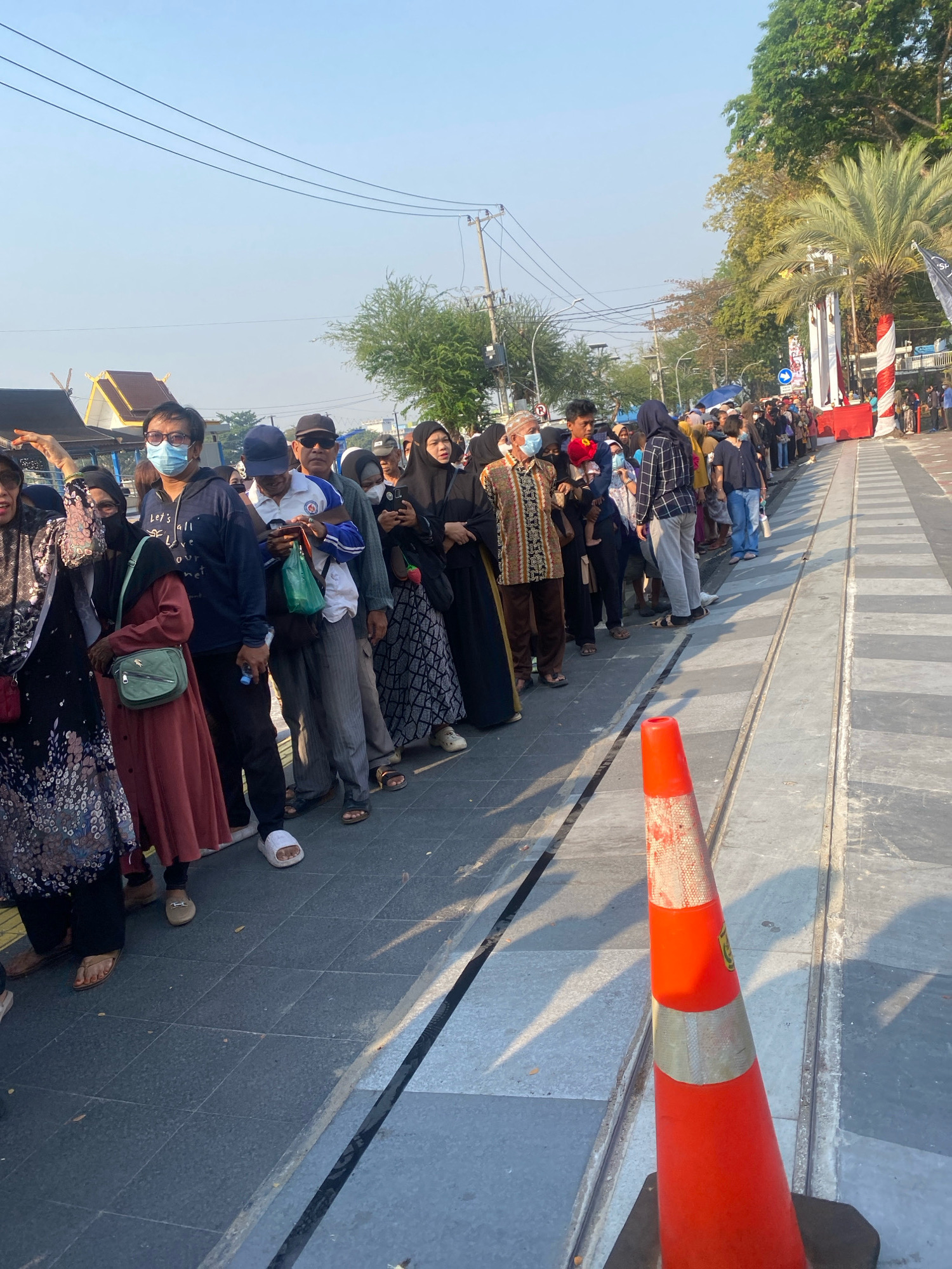 In Banjarmasin, Indonesia, a long queue of people stands on a sidewalk. The individuals are mostly dressed in modest attire, with many women wearing headscarves. The people are waiting in line, suggesting they are anticipating an event or service. The environment appears to be outdoors, with a clear sky and trees visible in the background. A bright orange traffic cone stands in the foreground, indicating a potential barrier or demarcation. The overall scene depicts a gathering of people in an urban setting.
