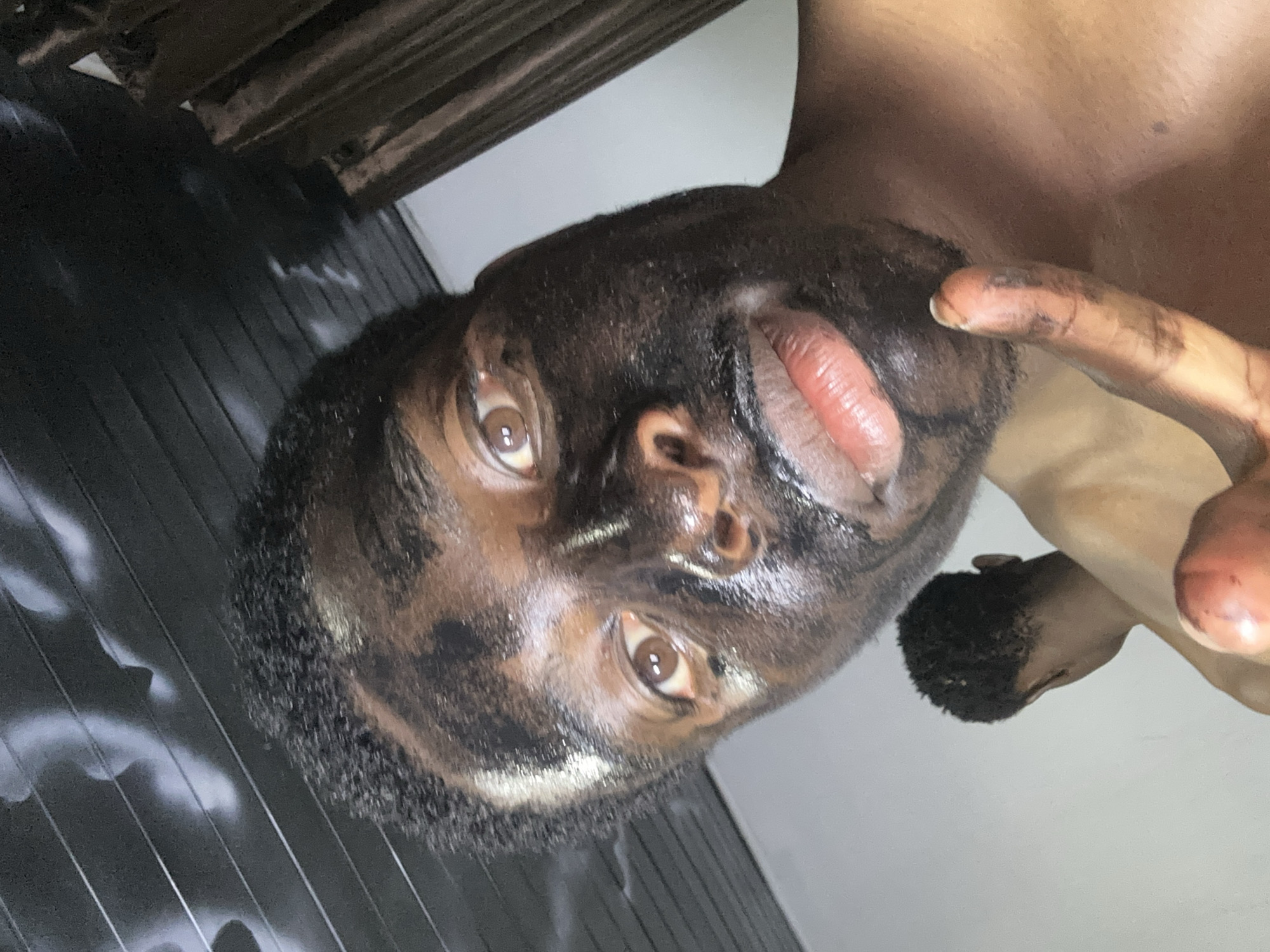 A close-up, angled view features a dark-skinned person with their face covered in a dark substance, possibly mud or clay. The person is looking upwards at the camera with their mouth slightly open. A finger, also covered in the dark substance, is visible near their mouth. The background shows a gray surface with white markings, possibly a wall or a floor, and part of what appears to be a wooden structure. There is no information present to indicate that the location is Bukuru, Nigeria.