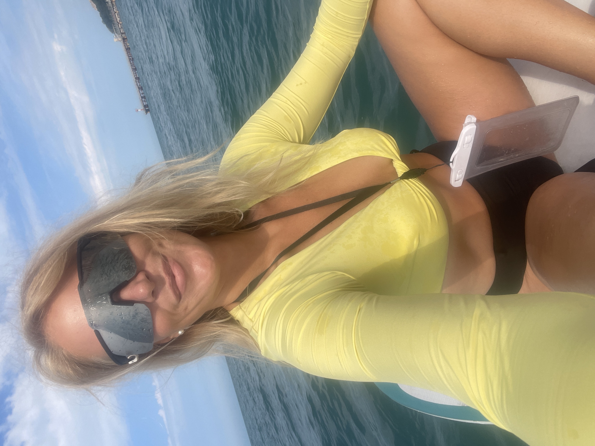 A woman with blonde hair is on a paddleboard in the Black Sea, with Sochi, Russia in the background. She is wearing a yellow long-sleeved rash guard, black bottoms, and large black sunglasses. She is holding a waterproof phone case around her neck. In the distance, a pier with a lighthouse extends into the sea, and buildings are visible on the shoreline. The sky is blue with some white clouds.