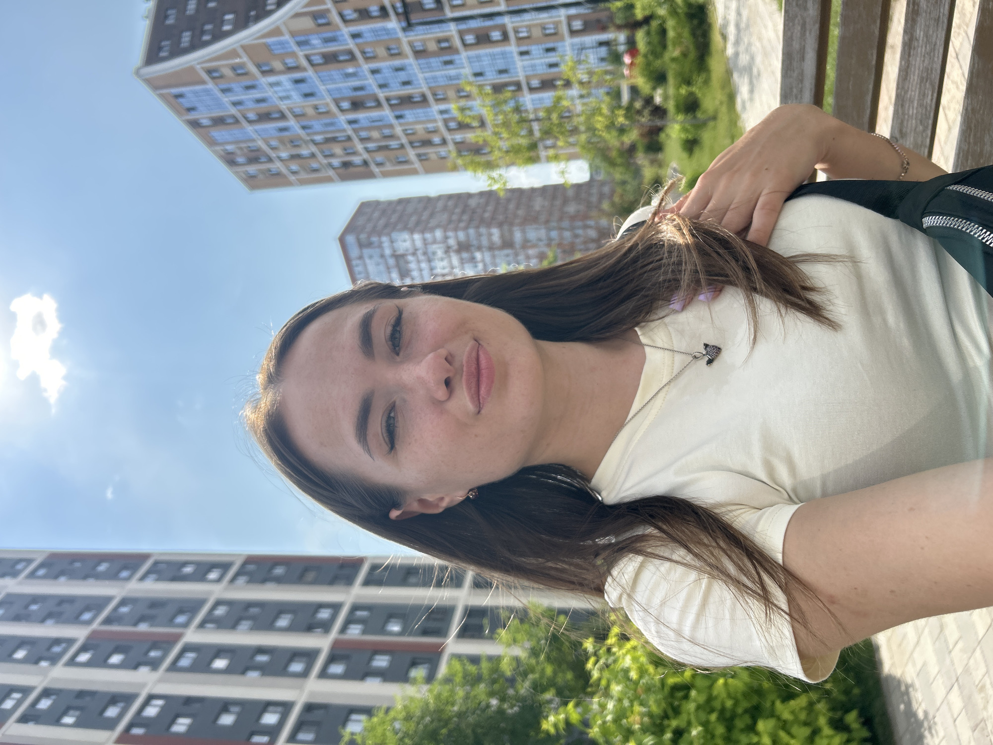 A young woman with long brown hair and fair skin is taking a selfie outdoors. She is wearing a light-colored t-shirt and a necklace with a pendant. Her hair is partially obscuring her face and shoulders. Behind her, several tall, modern apartment buildings are visible, along with lush green trees and a clear blue sky with a few clouds and bright sunlight. She appears to be sitting on a wooden bench.