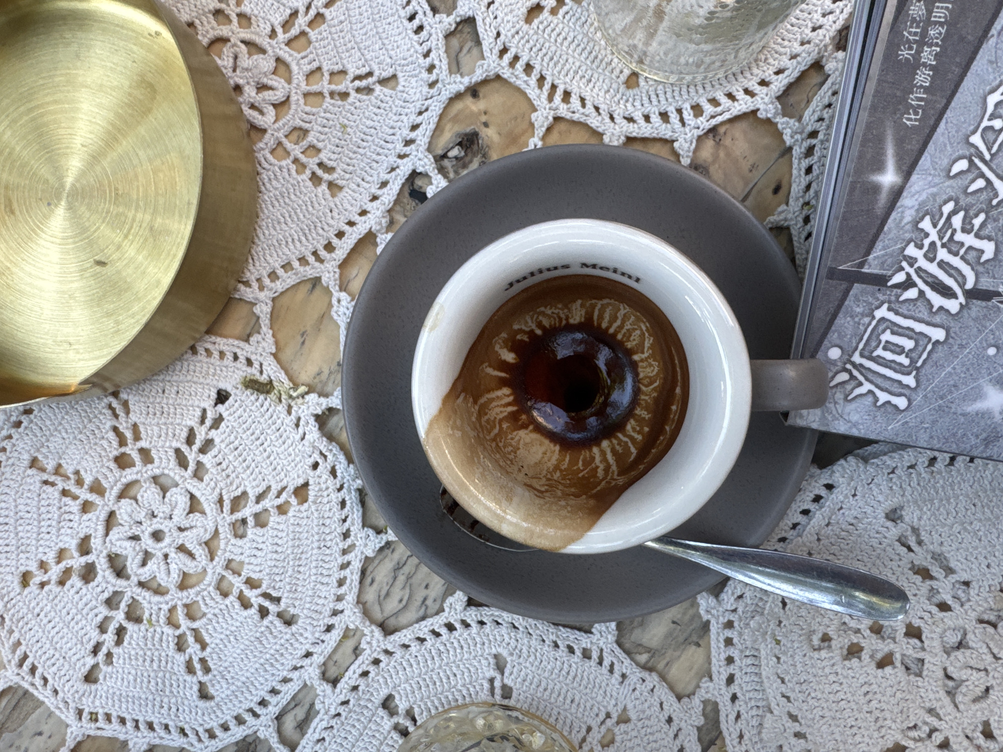 A top-down view showcases a coffee cup on a grey saucer, accompanied by a spoon, a golden dish, and a book with Asian characters, all resting on a white crocheted doily. The coffee cup appears to be a "Julius Meinl" branded cup and contains a coffee drink, such as espresso. The scene evokes a café setting, possibly in Belgrade, Serbia.