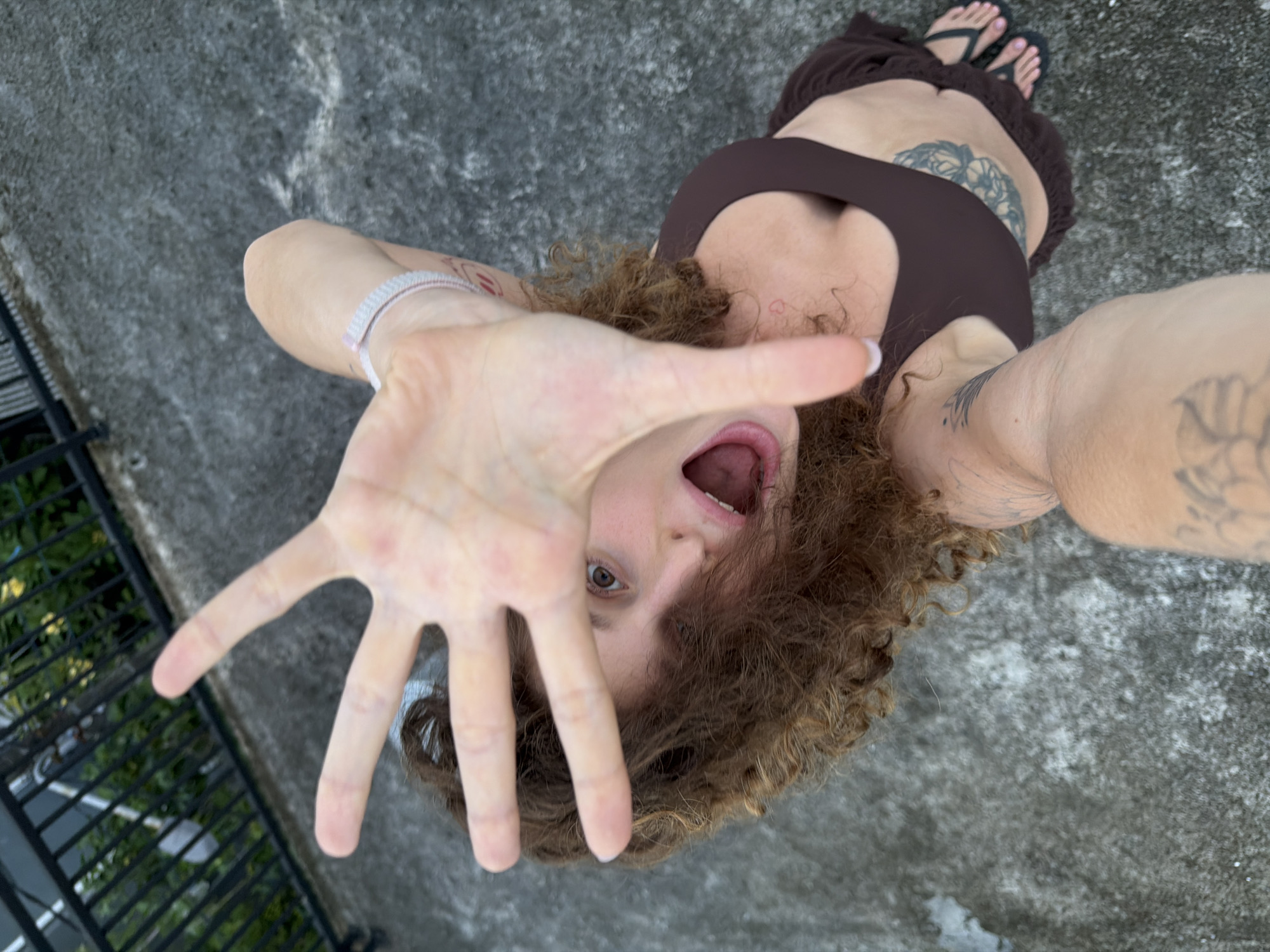 A woman is pictured from a low, upward-facing angle in North Kuta, Indonesia. She is wearing a brown top and a skirt with sandals and has multiple tattoos on her body. Her hair is curly. Her mouth is open, and her right hand is extended towards the camera. A black metal fence is visible on the left side of the frame, with green vegetation beyond it. The ground is grey concrete.