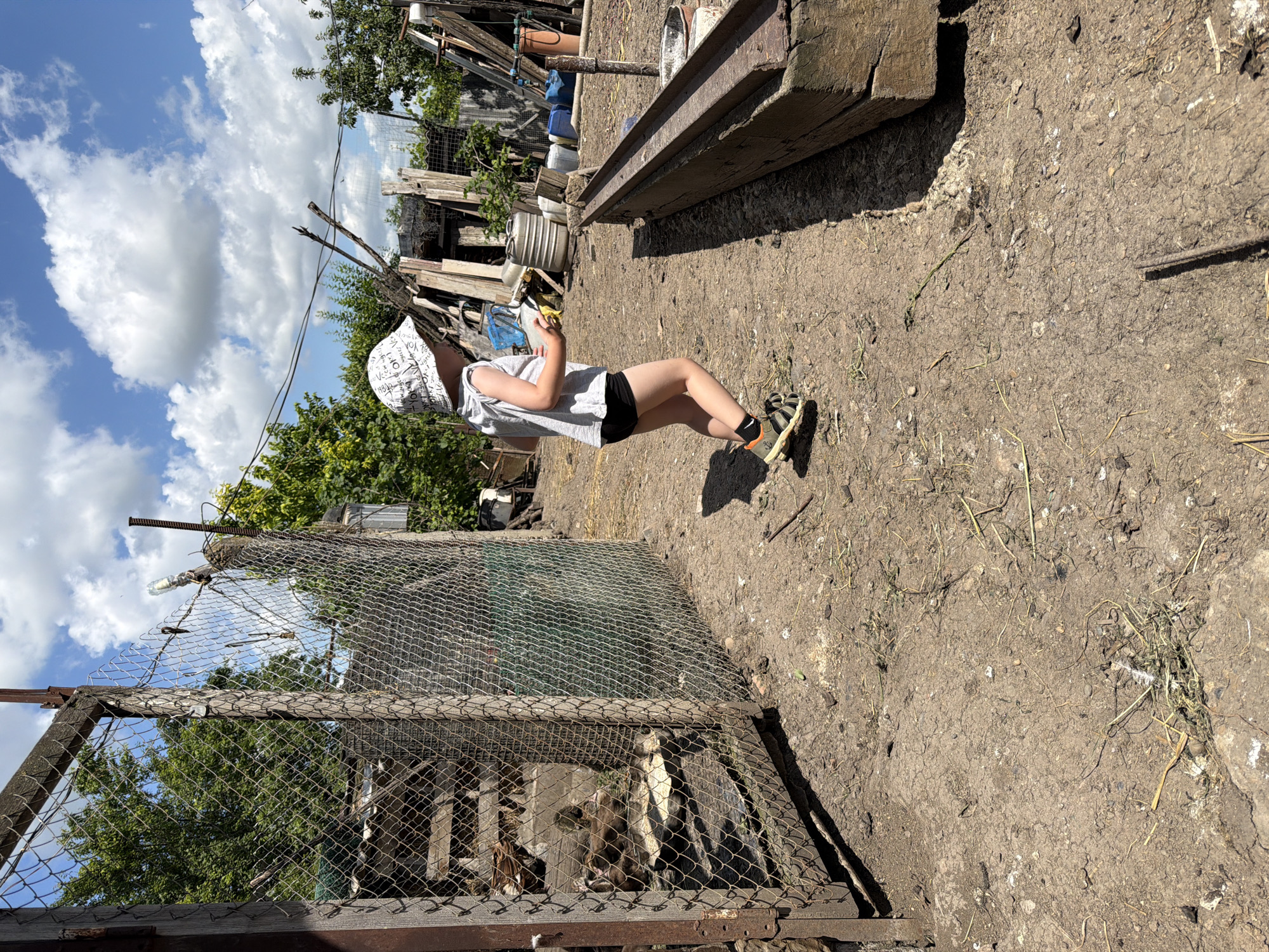 In Ust'-Dzhigutinka, Russia, a young child wearing a white bucket hat, a grey t-shirt, and black shorts is walking past a wire mesh enclosure on a sunny day. The enclosure, constructed with wooden posts, contains a few animals that appear to be goats. The ground is covered in dry dirt and scattered straw. To the right of the child, a wooden trough is visible. In the background, there are trees, various structures made of wood and metal, and large blue barrels. The sky is blue with scattered white clouds.