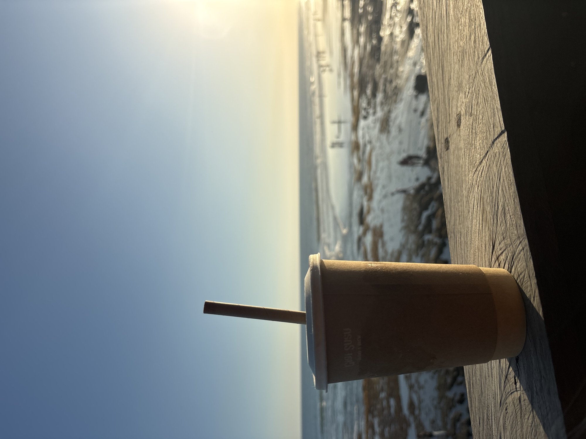 This media file shows a close-up of a brown disposable cup with a straw, resting on a wooden surface. The cup has white text on it that reads "GUL SUSU". Behind the cup, a body of water with ice or snow is visible, with the sun low in the sky creating a bright, hazy effect. The sky is a clear blue. The overall scene suggests an outdoor setting, possibly a beach or a waterfront, during a cold time of day or year, despite the location context of North Kuta, Indonesia. The bright sun and clear sky could indicate a cold but sunny day. The wooden surface appears weathered, with visible grain and some scratches. The image is vertically oriented.