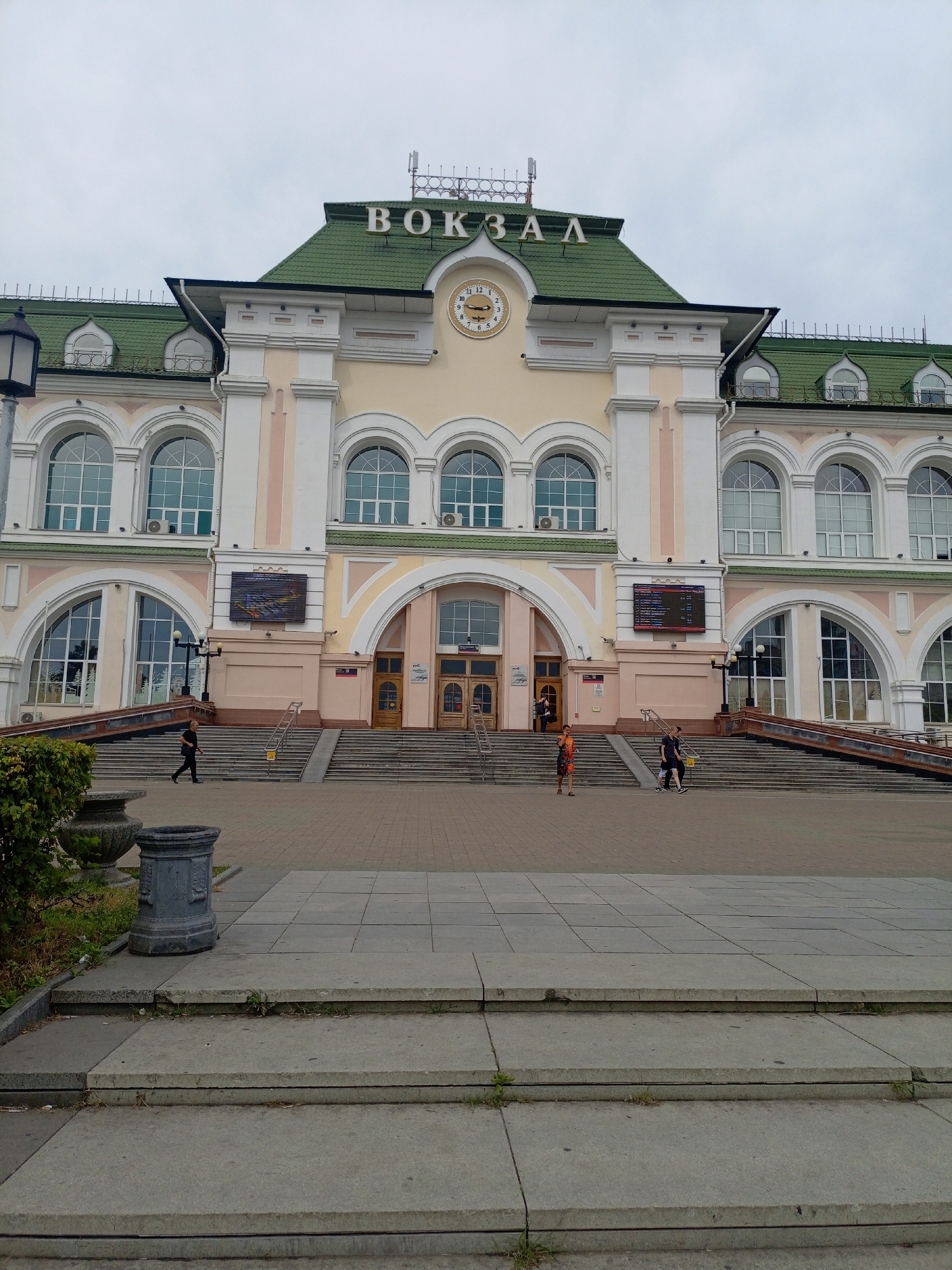 The image shows the exterior of a train station building with "ВОКЗАЛ" written in large white letters across the top. The building has a light yellow facade, white columns, and a green pitched roof. There are arched windows and a large clock on the front of the building. A set of wide concrete stairs leads up to the main entrance, with ramps on either side. Several people are visible on the stairs and in the plaza in front of the building. A lamppost is visible on the left side of the stairs. Two large stone planters are located to the left of the stairs, one containing a dark grey trash receptacle. The sky is overcast.