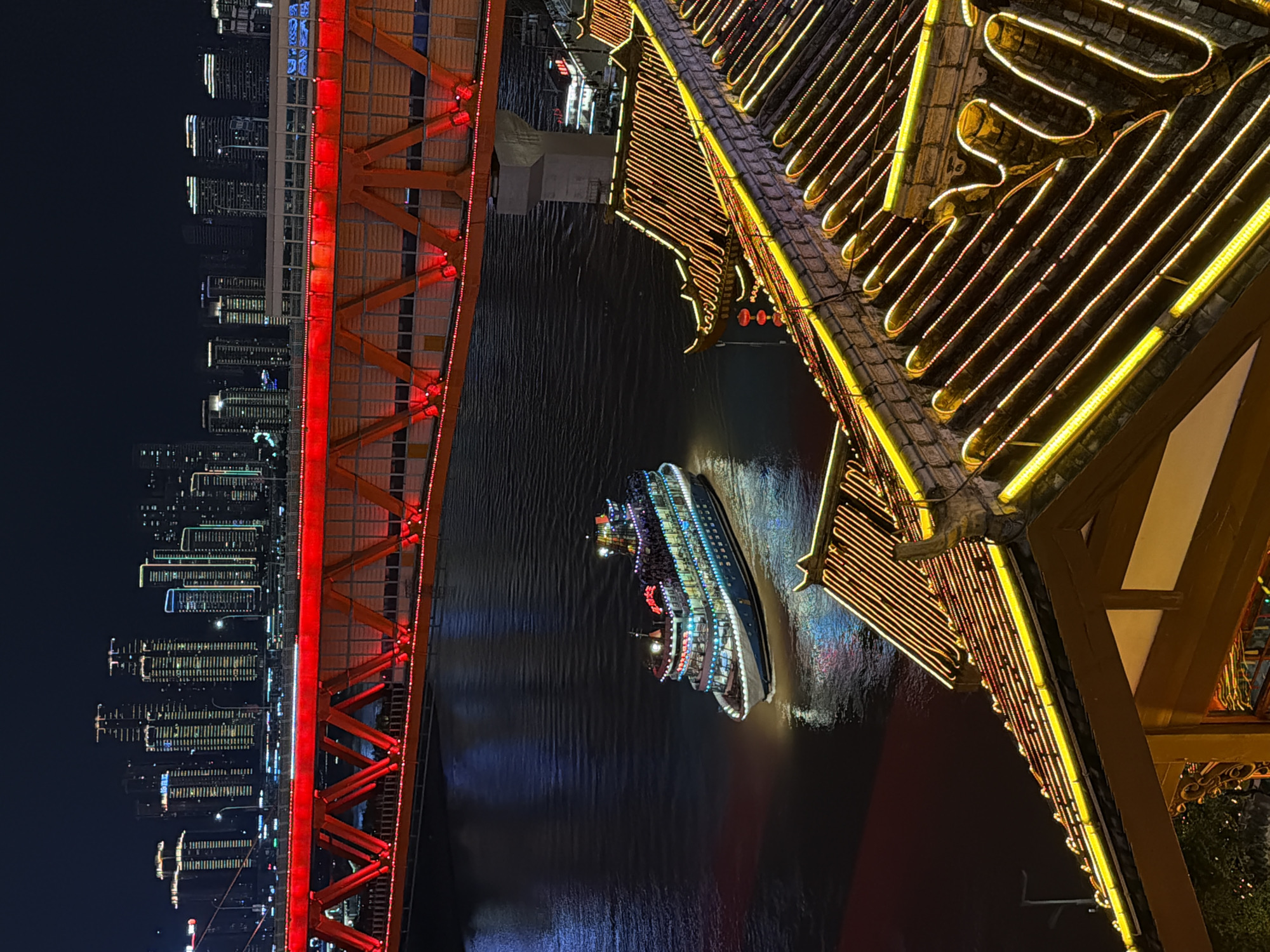 The image depicts a nighttime scene in the Yuzhong District of Chongqing, China. A brightly lit passenger boat navigates the dark waters of a river, reflecting the colorful lights of the city. In the foreground, traditional Chinese-style rooftops adorned with yellow and white LED lights are visible. A large, illuminated red bridge spans the river in the mid-ground, with the illuminated skyline of modern skyscrapers forming the backdrop. The scene is illuminated by artificial lights from the city, the bridge, the boat, and the decorative lighting on the buildings.
