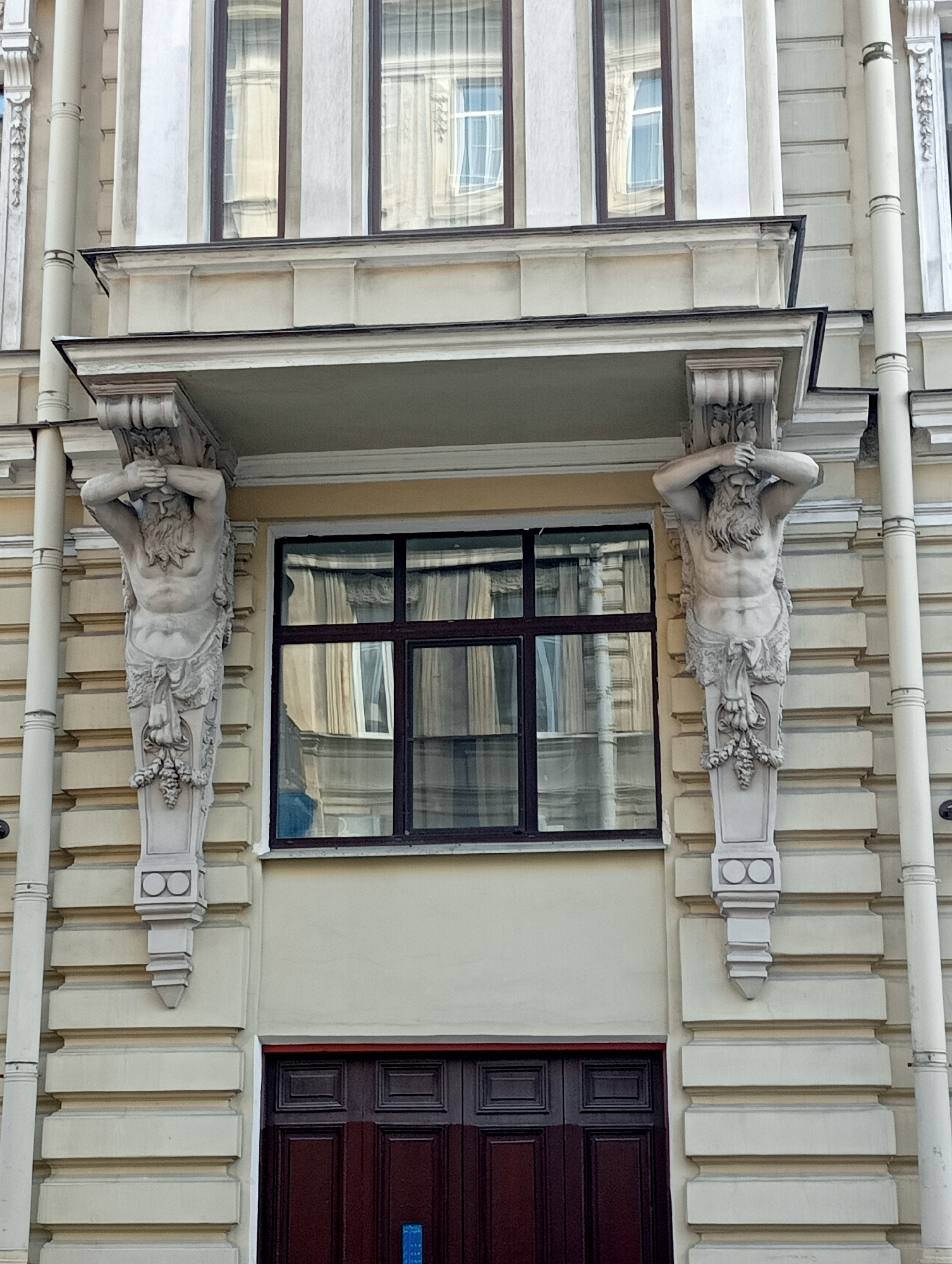 The image shows the facade of a building, likely in Saint Petersburg, Russia, featuring ornate architectural details. Two muscular statues, appearing to be atlantes or caryatids, are positioned on either side of a large window. These figures are carved in stone, with one on the left and another on the right, each supporting the structure above them with their hands on their heads. They are depicted with beards and wear decorative draped clothing around their waists.
Below the statues, a dark, double-door entrance is visible, with a dark red-brown color and panel detailing. The building's exterior is characterized by a light beige color, with horizontal and vertical decorative elements, including pilasters and layered cornices. The window above the doors is a large, multi-paned structure with dark framing, reflecting the surrounding architecture and sky. A decorative pipe runs vertically along the right side of the facade. The overall impression is one of classical architectural grandeur.