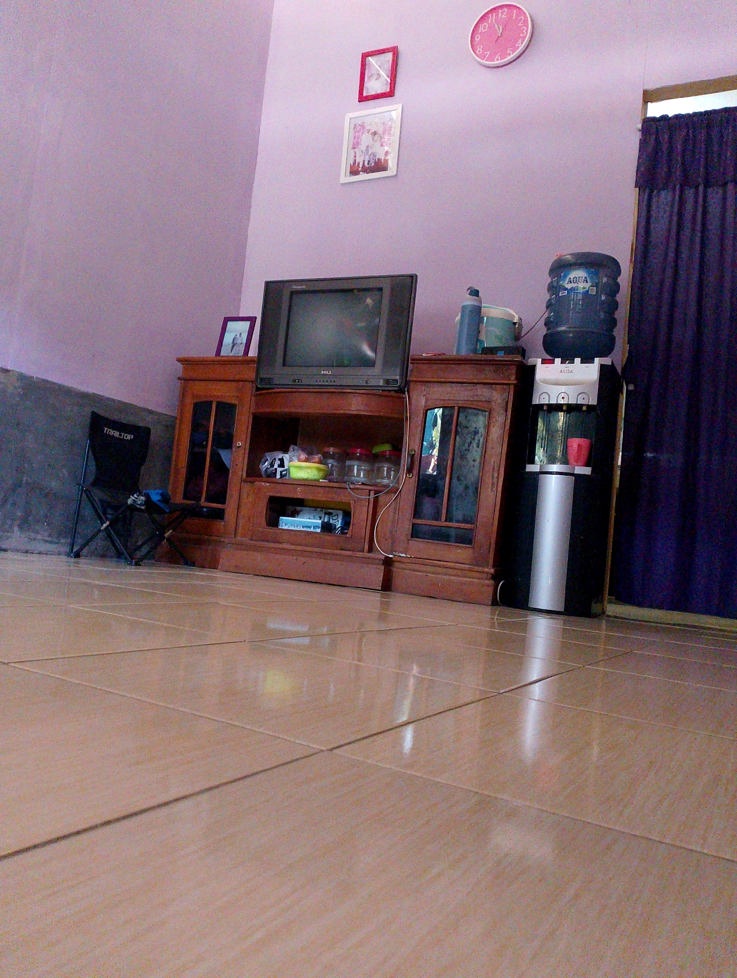 This image depicts an indoor scene, likely a living room or common area, within a dwelling in Desa Babakan, Indonesia. The foreground is dominated by a tiled floor, reflecting light and showing the texture of the tiles. In the mid-ground, a wooden entertainment center stands against a pale lavender wall. On top of the entertainment center, a vintage television set is positioned centrally, with various items placed around it, including jars containing food items and a small picture frame. To the right of the entertainment center, a black and silver water dispenser is situated, topped with a large blue water bottle labeled "AQUA". A red cup is visible within the dispenser's dispensing area. To the left of the entertainment center, a black foldable chair with the text "TRAILTOP" is visible. In the background, a dark purple curtain hangs on the right side of the frame, partially obscuring a window or doorway. On the wall above the entertainment center, a pink analog clock with Roman numerals is mounted, alongside two framed pictures. The overall impression is of a lived-in space with a functional arrangement of furniture and appliances.