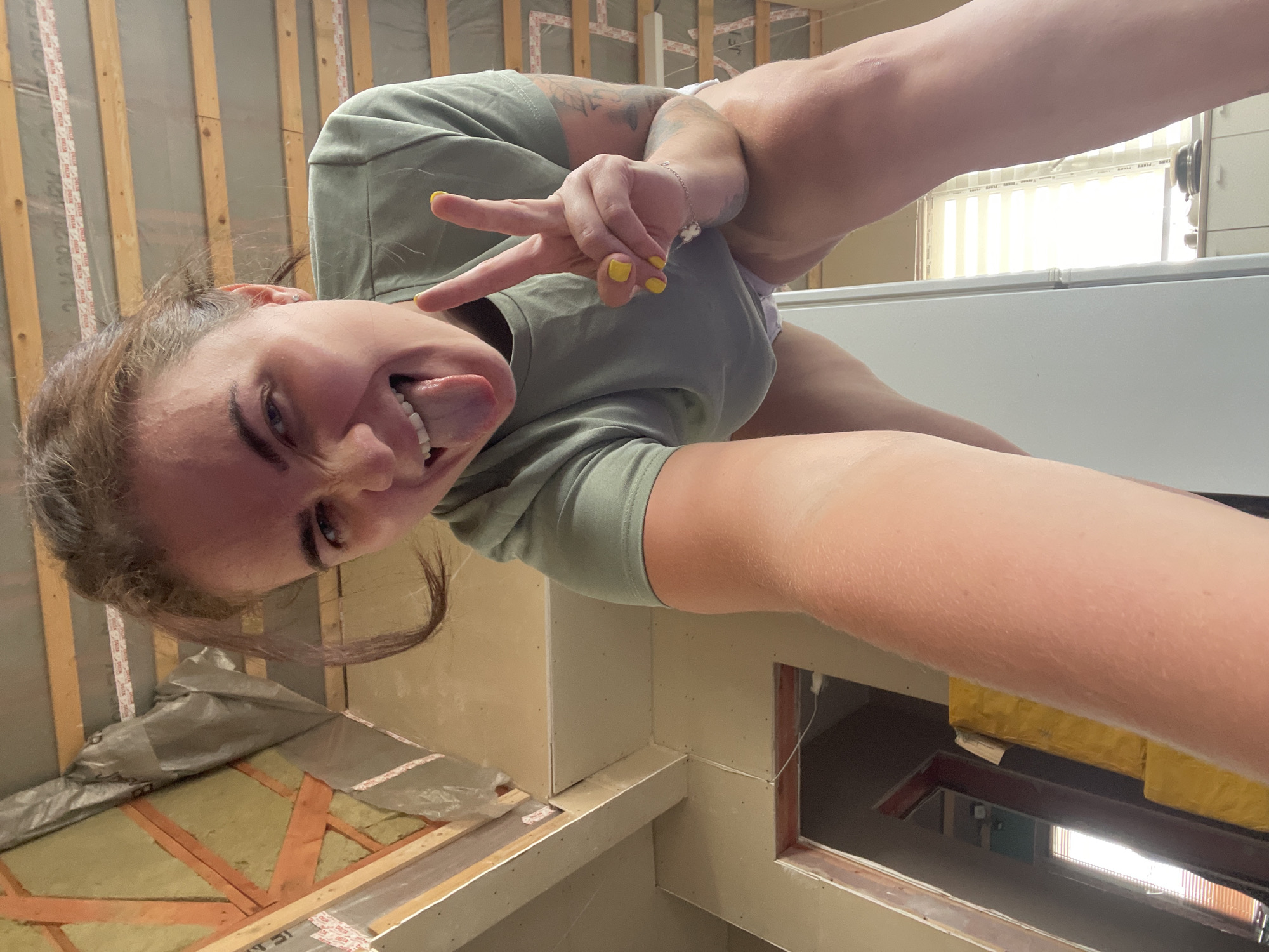 In this media file, a young woman with brown hair is captured in a selfie. She is wearing a light green t-shirt and has her tongue sticking out, with her right hand held up in a peace sign. Her nails are painted yellow. Tattoos are visible on her left arm. The background suggests a construction or renovation site, with wooden studs and insulation visible on the wall. The overall impression is playful and energetic.
The image is oriented vertically and appears to have been taken indoors, possibly during the day, given the bright light coming from a window in the background. The woman's expression and pose convey a sense of fun and lightheartedness. The setting includes construction materials, suggesting an ongoing project. There is text visible on some of the construction tape, but it is not clearly legible. The location context provided is "Мосметаллоконструкция Онт, Russia."