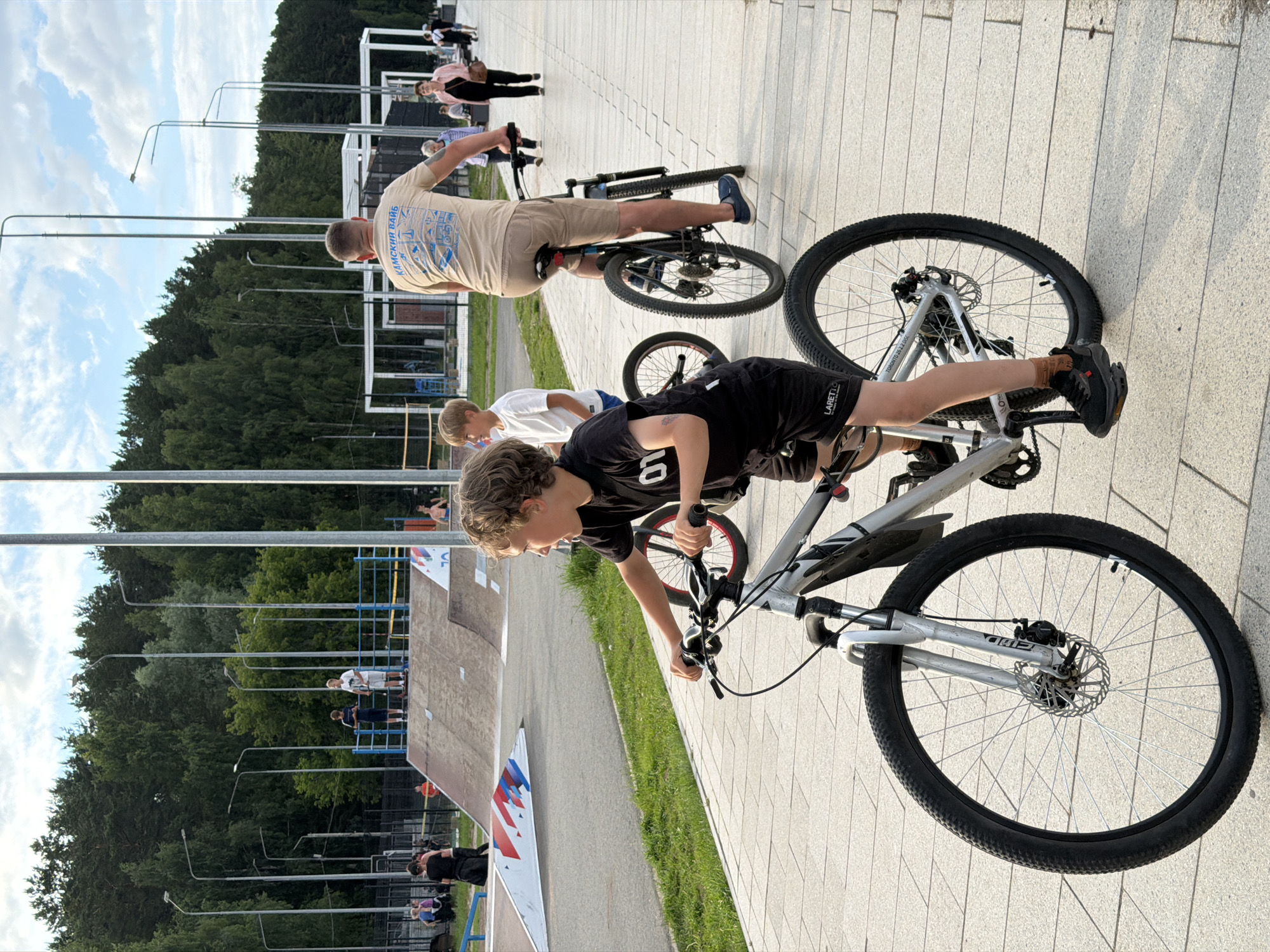 In Chaikovsky, Russia, a boy with curly brown hair rides a silver mountain bike on a paved walkway. He is wearing a black t-shirt and shorts. His bike has disc brakes on the front wheel and a chain guard. In the background, a man on a bicycle wears a light brown t-shirt with Russian text and tan shorts. Behind him, several people walk along the path, and a skate park with ramps is visible. Tall trees line the edge of the park, and the sky is partly cloudy.