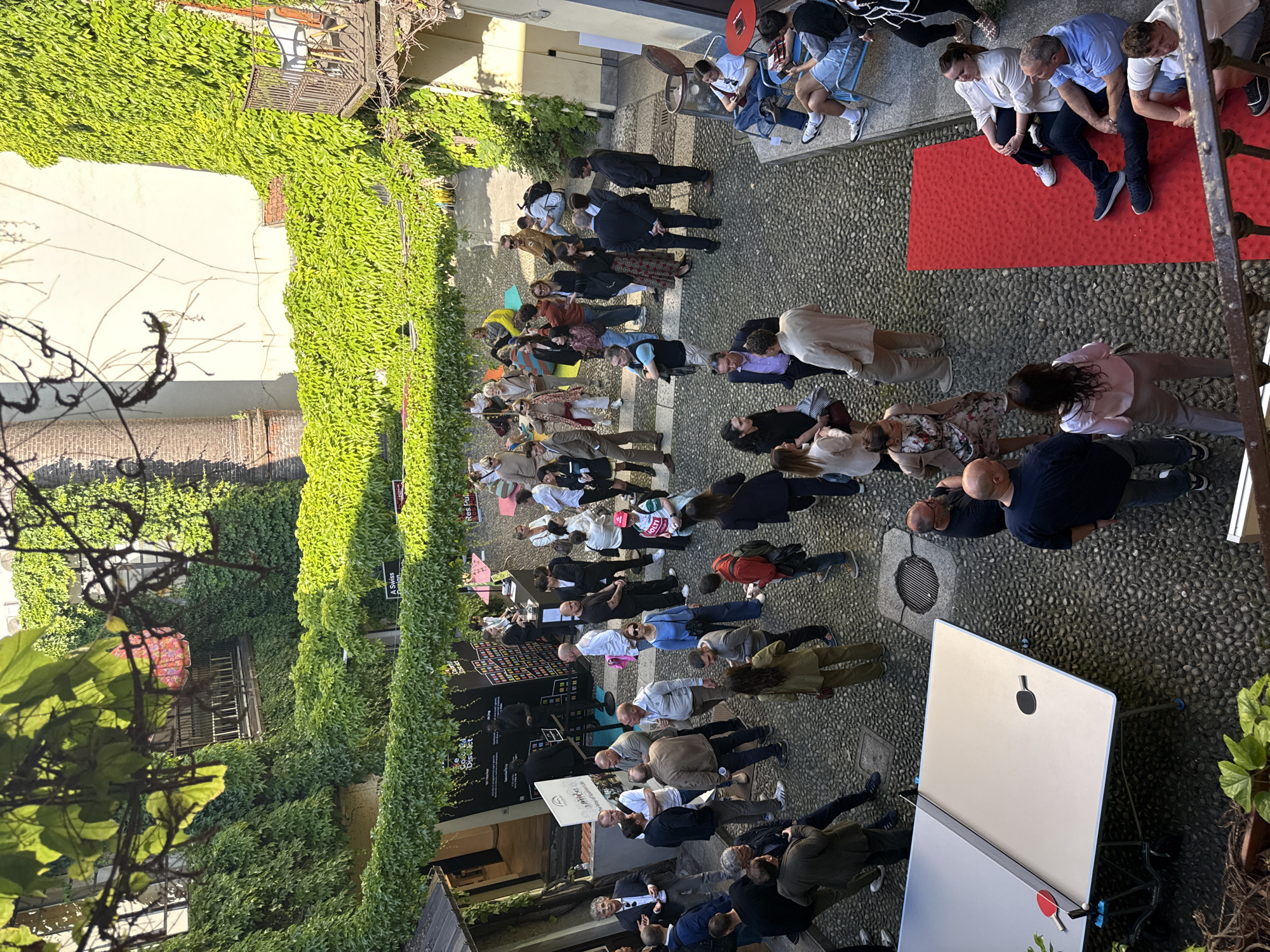 This image captures an outdoor gathering in a sunlit cobblestone courtyard, likely in Milan, Italy, given the context. The scene is bustling with a large group of people, primarily adults, standing and engaging in conversations. Most individuals are dressed in business casual or smart casual attire, suggesting a professional or semi-formal event.
The courtyard is enclosed by buildings with walls extensively covered in vibrant green ivy, creating a natural and inviting atmosphere. Bright daylight indicates good weather, casting clear shadows on the ground and people.
In the upper left portion of the image (when oriented correctly), a few people are seated at small tables with blue chairs and a red table. Throughout the courtyard, individuals are clustered in small groups, interacting. Towards the right side, two distinct red mats or carpets are placed on the cobblestones.
Notable objects include a large, reflective display screen or sign with a silver finish, featuring a stylized black paddle-like logo (resembling an Apple logo) on its surface. Near this screen, a well grate is visible on the ground. On the left side of the frame, several tall, black banners with white text are visible. Visible text fragments include "Res Publica", "A Swiss...", and "District", likely indicating sponsors or event themes. One person holds a white sign that clearly reads "Sustainability by Design". Another person carries a pink tote bag with partially visible text, possibly "H_E__Y". The overall mood appears relaxed and social, with attendees appearing engaged in the event.