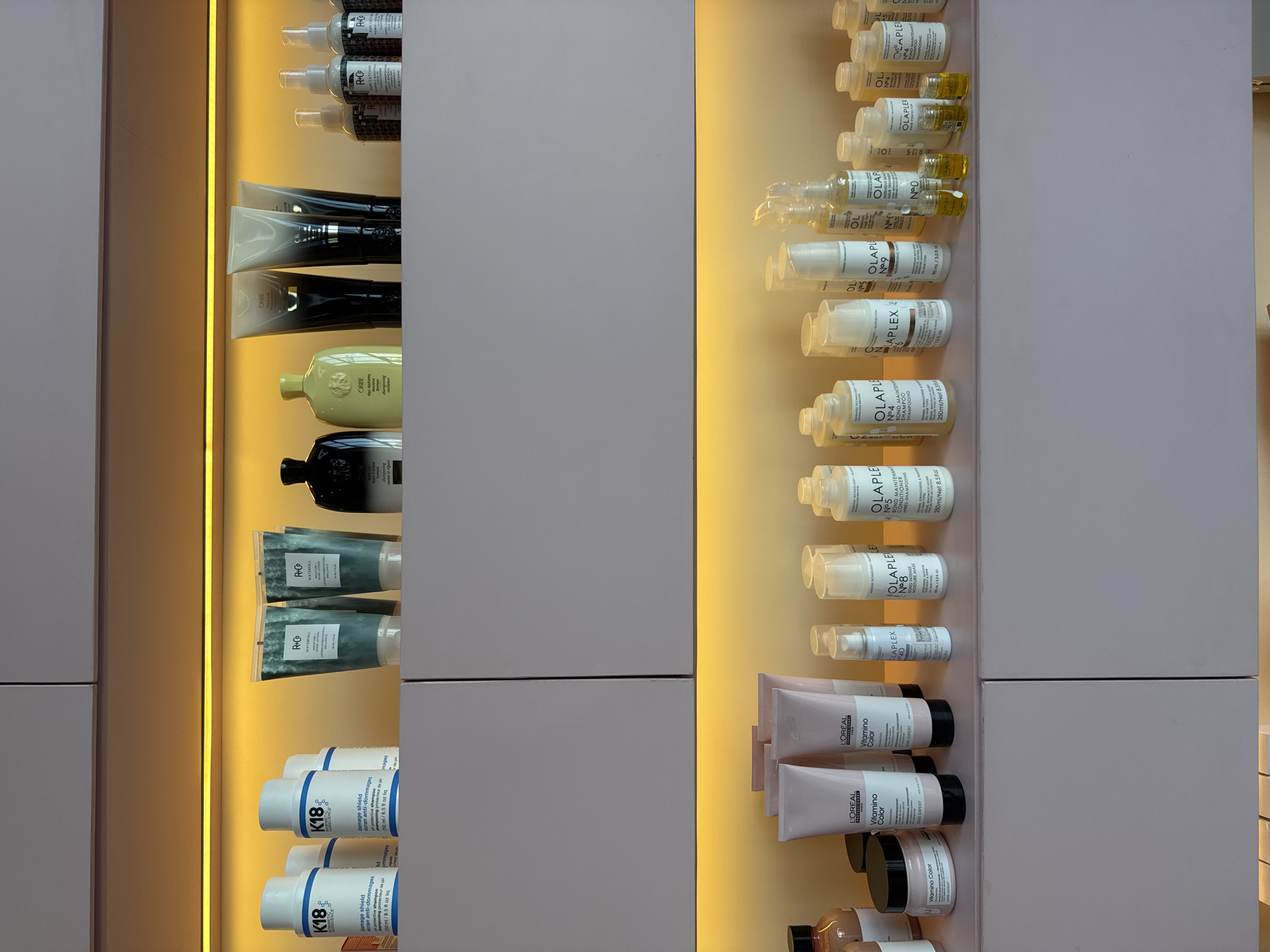 This is a vertical, eye-level shot of a retail display case for hair care products. The display case is illuminated by warm yellow LED lighting, creating a soft glow. The products are arranged on shelves made of a pale pink material.
On the left side of the display, there are rows of various hair products. These include tubes and bottles of shampoo, conditioner, and other treatments. Some of the visible brands include R+Co, Oribe, and K18. The K18 products are white bottles with blue and silver branding, and text like "damage shield" and "cran anti-dommages" is visible.
The right side of the display features a tiered arrangement of smaller bottles, primarily from the OLAPLEX brand. These are mostly clear bottles with white labels, and the text "OLAPLEX," "No. 4," and "No. 8" are prominent. Some of these bottles contain yellow liquid. Further down on the right are pink tubes of L'Oréal hair products, with "Vitamino Color" and "THE EXPERT" visible.
The overall impression is one of a clean, well-organized, and modern retail environment, likely a salon or beauty store. There are no people or animals visible in the frame. The focus is entirely on the presentation of the hair care products. The lighting suggests it could be evening or an indoor setting with artificial light. The scene is static, with no activity taking place other than the passive display of goods.