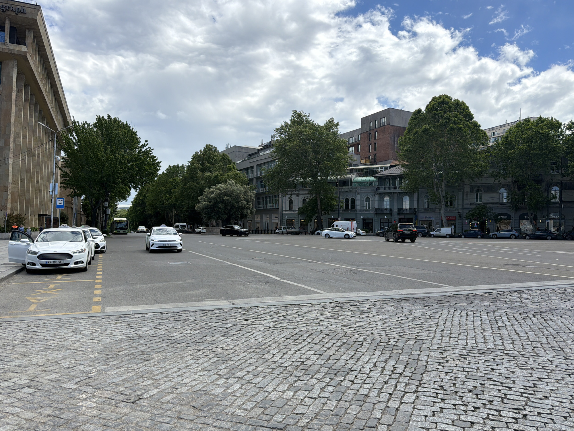 An outdoor wide-angle shot captures a vibrant urban street or square under a partly cloudy sky, indicating daytime. The immediate foreground features a textured cobbled pavement, which transitions into a broad asphalt road designed for multiple lanes of traffic.
On the left side of the road, a designated taxi stand is marked by yellow painted lines and a prominent "TAXI" stencil on the asphalt. Several white sedans, notably two with "TAXI" signs on their roofs, are parked here. One white taxi has its front driver's side door open, and its license plate "KK-305-SK" is clearly visible, suggesting a location in Georgia. A blue square sign with a white car and the word "TAXI" is mounted on a pole nearby. Indistinct figures of people can be seen near the parked vehicles and sidewalks. Further down the road, a green bus and other cars, including a black SUV and a white van, are either driving or parked.
The street is flanked by tall, leafy green trees and a mix of buildings. On the left, a large, multi-story building with a distinct, ribbed vertical facade shows the partial text "graph" on its upper section. On the right, the architecture varies, featuring buildings with traditional arched windows, some with modern elements, and one with a notable glass-covered section. Faint text on storefronts in the distance includes "GRAPE," "TIN," "OnaN," "ACCOR," and "XRR." The scene depicts a typical busy day in a city, with various vehicles and everyday urban activity.