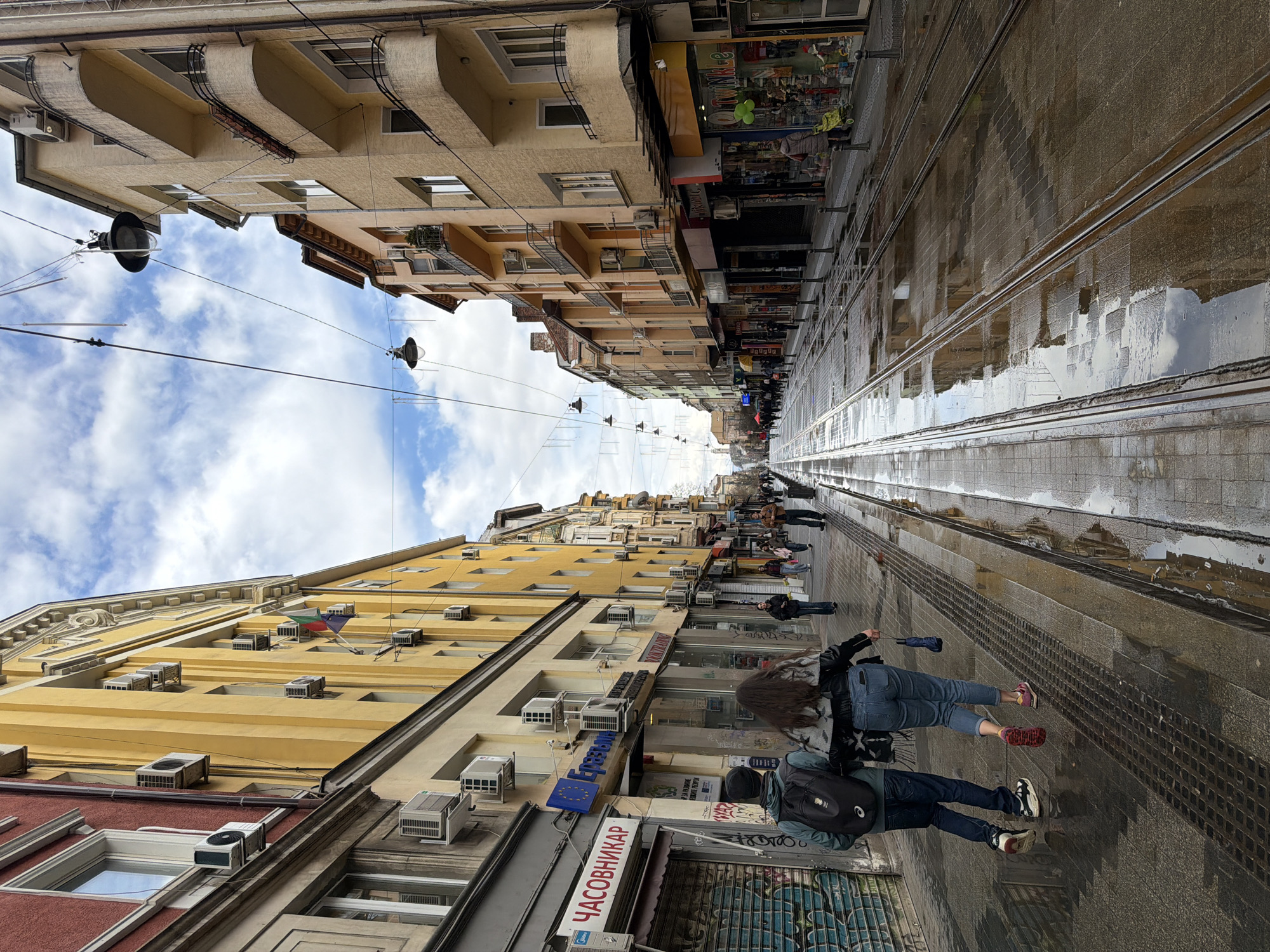 The image shows a street in Sofia, Bulgaria on a wet day. The view is down a pedestrianized area with tram tracks running through the center. Buildings line both sides of the street, varying in height and architectural style, mostly in shades of yellow and beige. There are multiple air conditioning units visible on the facades.
People are walking along the street. Some shops are visible on the sides. Overhead, there are decorative lights strung across the street. The street surface is wet, with puddles reflecting the sky. The sky is cloudy with patches of blue. There is a sign in Cyrillic on one of the buildings.