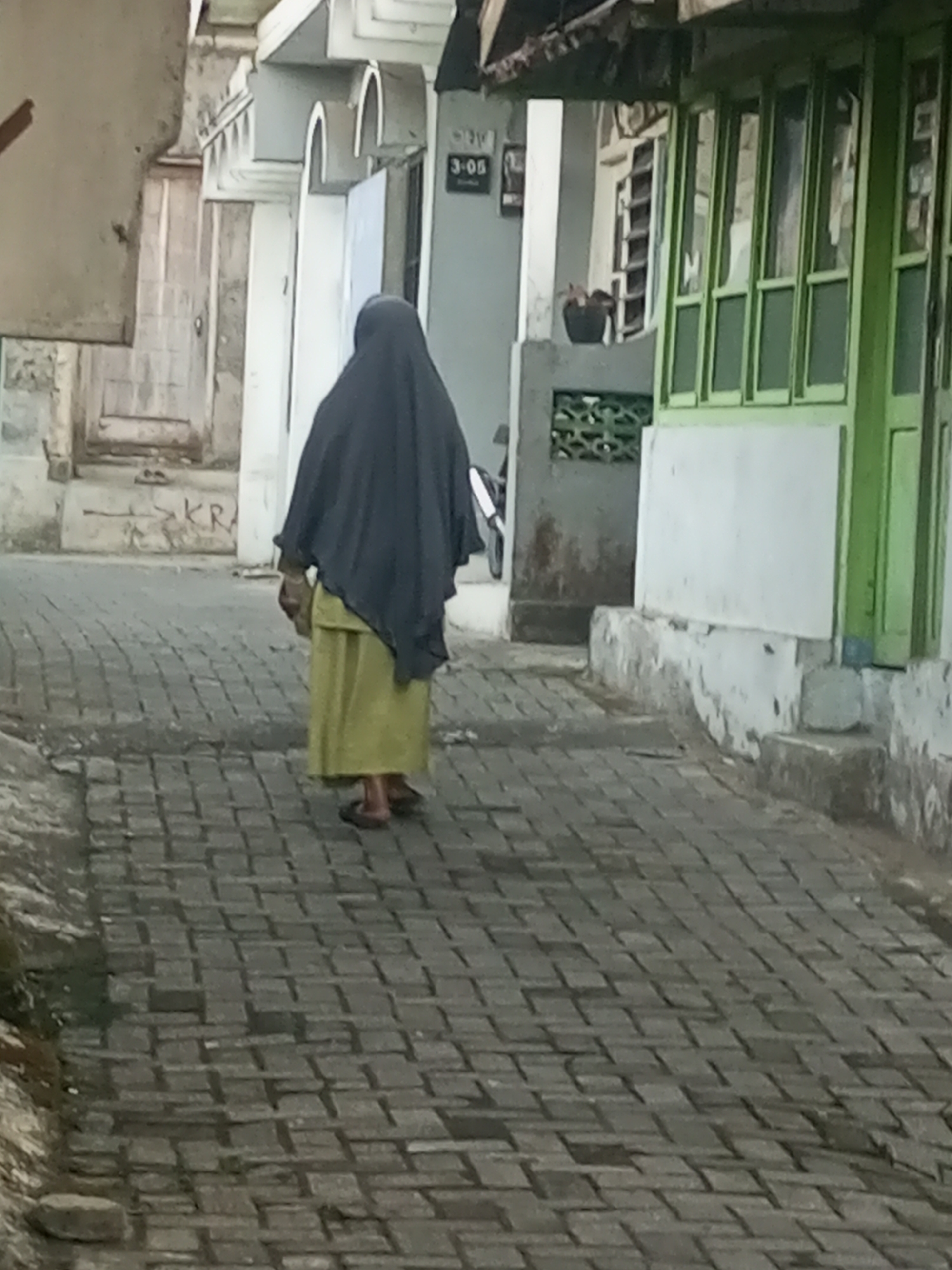 An individual wearing a dark headscarf and a light green skirt walks away from the camera on a brick-paved pathway in Andongsili, Indonesia. The pathway runs between buildings on either side. The building to the left appears to be older, with a light-colored facade and some graffiti. The building to the right has a vibrant green facade with multiple windows. The scene appears to be an outdoor walkway or street in a residential or commercial area.