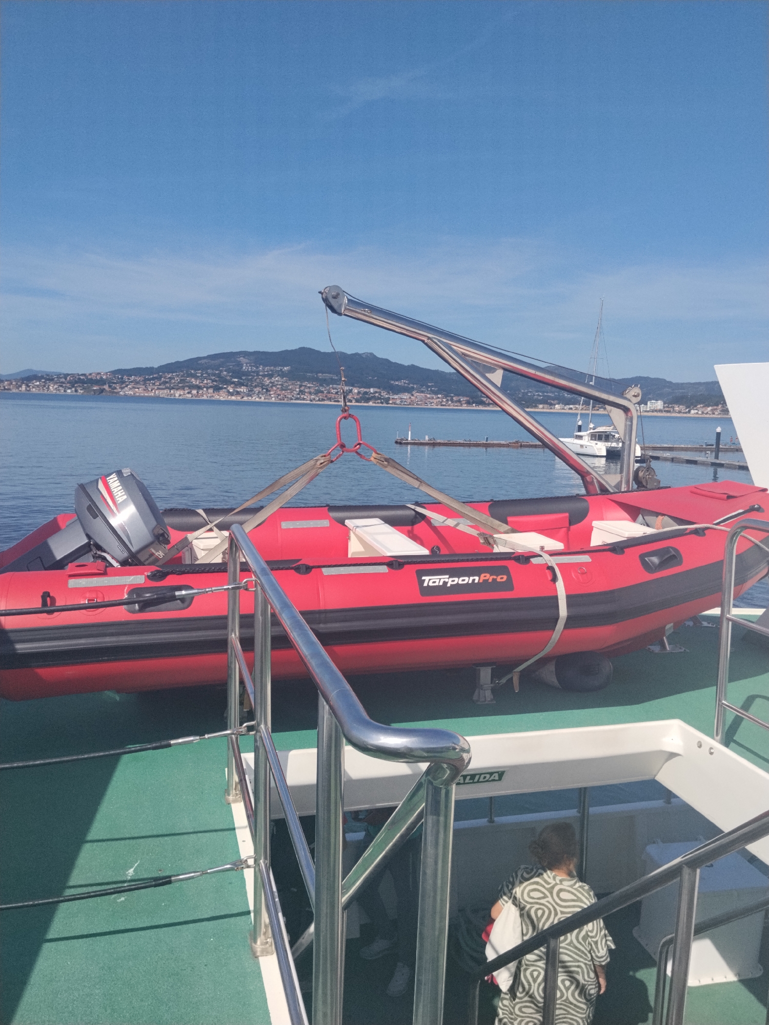 A red and black inflatable boat, identified by the "TarponPro" logo, is suspended by a crane over the deck of a larger vessel. The boat has a Yamaha outboard motor. In the background, the bay of Baiona, Spain, is visible with a town nestled on the hillsides and a breakwater extending into the water. A white sailboat is moored in the bay. On the deck of the larger vessel, a person wearing a patterned shirt and carrying a white bag walks down a set of stairs. The deck is painted green, and there are metal railings. The sky is clear and blue.