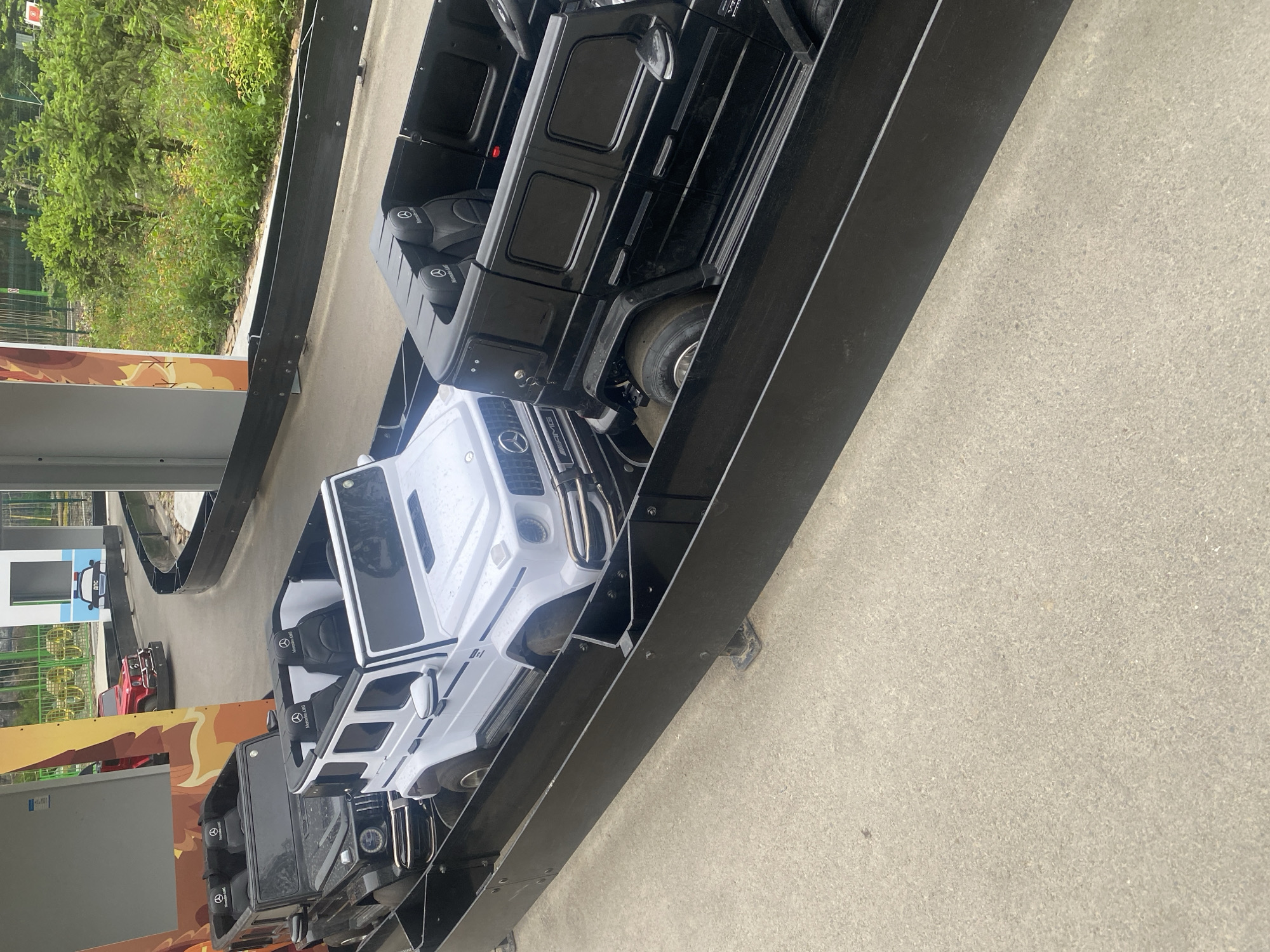 The image shows a go-kart track with several toy cars designed to resemble Mercedes-Benz G-Class vehicles. In the foreground, two open-top G-Wagon replicas are visible, one white and one black. Both have black interior seats with the Mercedes-Benz logo. Behind the white car, another black G-Wagon replica is partially visible. Further down the track, a red toy car, also resembling a G-Wagon, is seen. The track is bordered by black barriers. The environment appears to be outdoors, with greenery and trees in the background. The ground is paved with asphalt or concrete. The overall scene suggests a children's amusement park or a similar recreational facility.