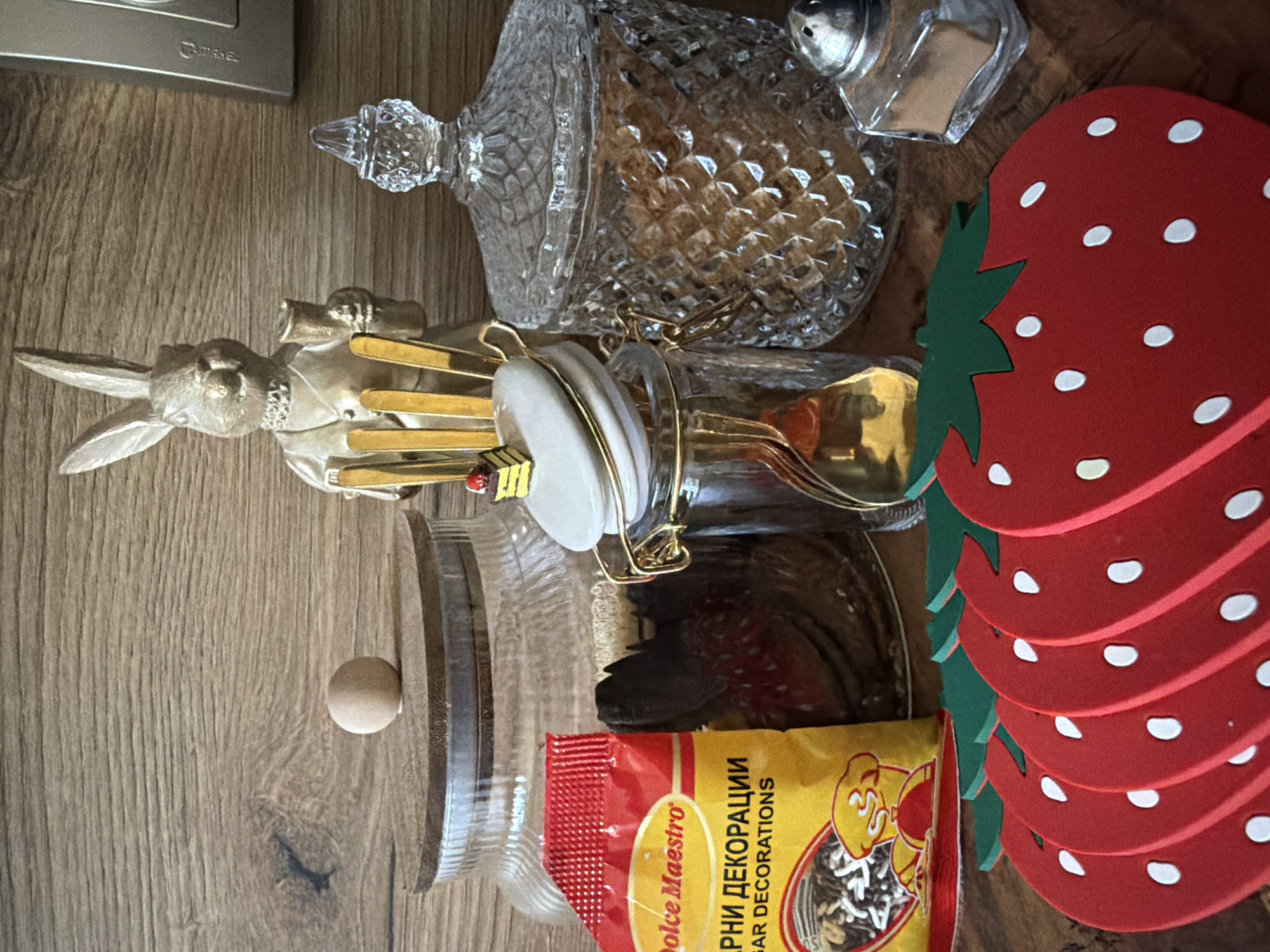 A still life image features various decorative and functional kitchen items arranged on a wooden surface. In the background, a wooden wall with a prominent grain pattern serves as the backdrop. To the left, a large glass jar with a wooden lid and a small, spherical knob sits partially filled with a light brown granular substance, likely sugar or flour. Next to it is a yellow packet of "Dolce Maestro" sugar decorations.
Dominating the center of the image is a decorative ceramic rabbit figurine dressed in a suit and holding a small vessel. In front of the rabbit, a small glass jar with a hinged lid contains gold-colored spoons. A tiny replica of a layered cake with a red cherry on top is affixed to the lid of this jar. To the right, a tall, ornate crystal jar with a lid is filled with more granular brown material. Adjacent to this crystal jar is a small, clear glass salt shaker with a metallic top.
In the foreground, a row of five red strawberry-shaped coasters, each with green leafy tops and white dot "seeds," are laid out. The scene appears to be indoors, possibly on a kitchen counter or shelf. The image does not contain any geographical markers or context to suggest a location within Pazardzhik, Bulgaria.