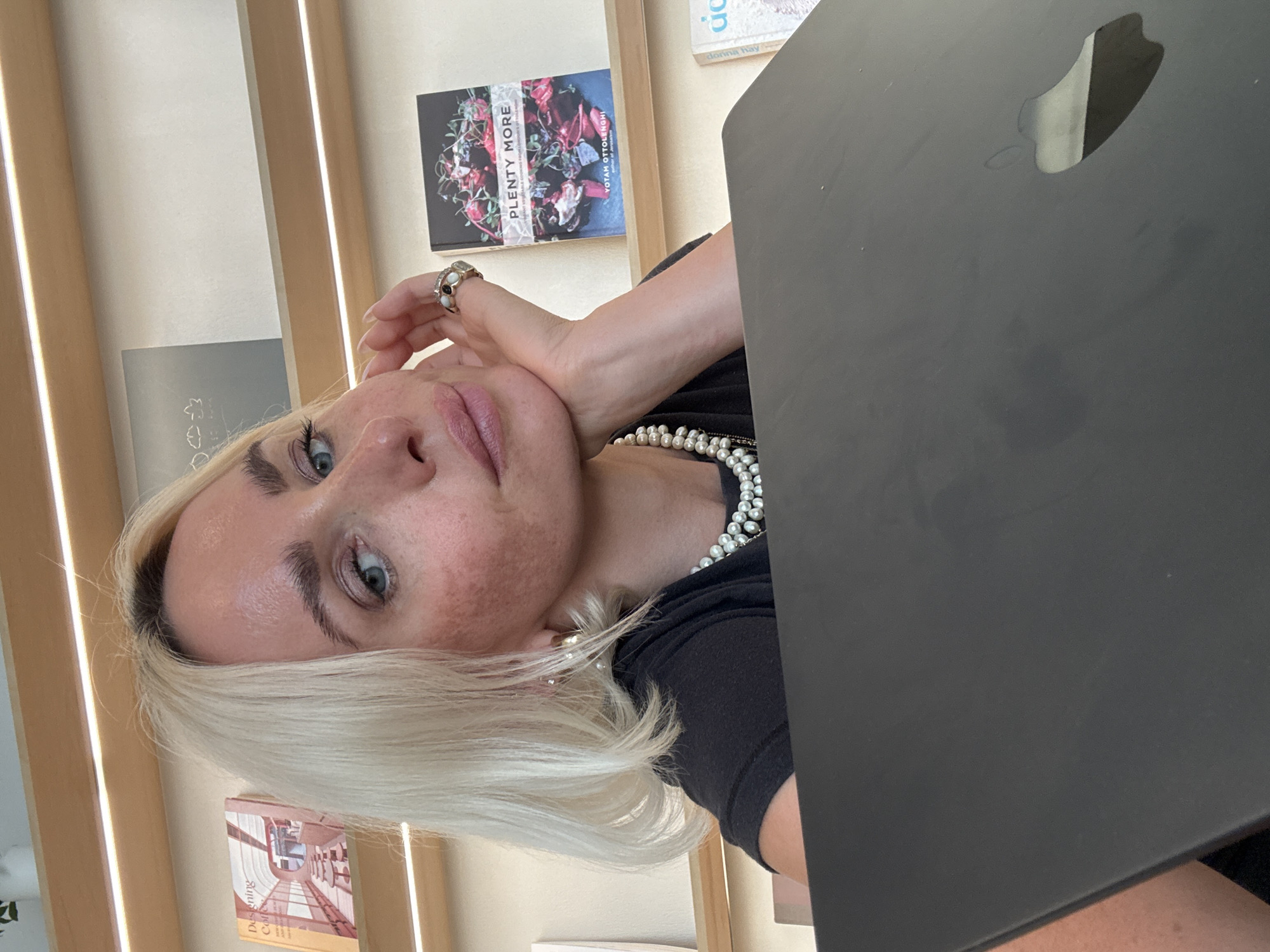 A blonde woman is captured in a selfie, holding her chin with her right hand. She is wearing a black t-shirt and a pearl necklace. The background consists of bookshelves filled with books, including one titled "Plenty More." The woman has light blue eyes and is looking at the camera. The lighting in the scene is bright, suggesting an indoor setting.