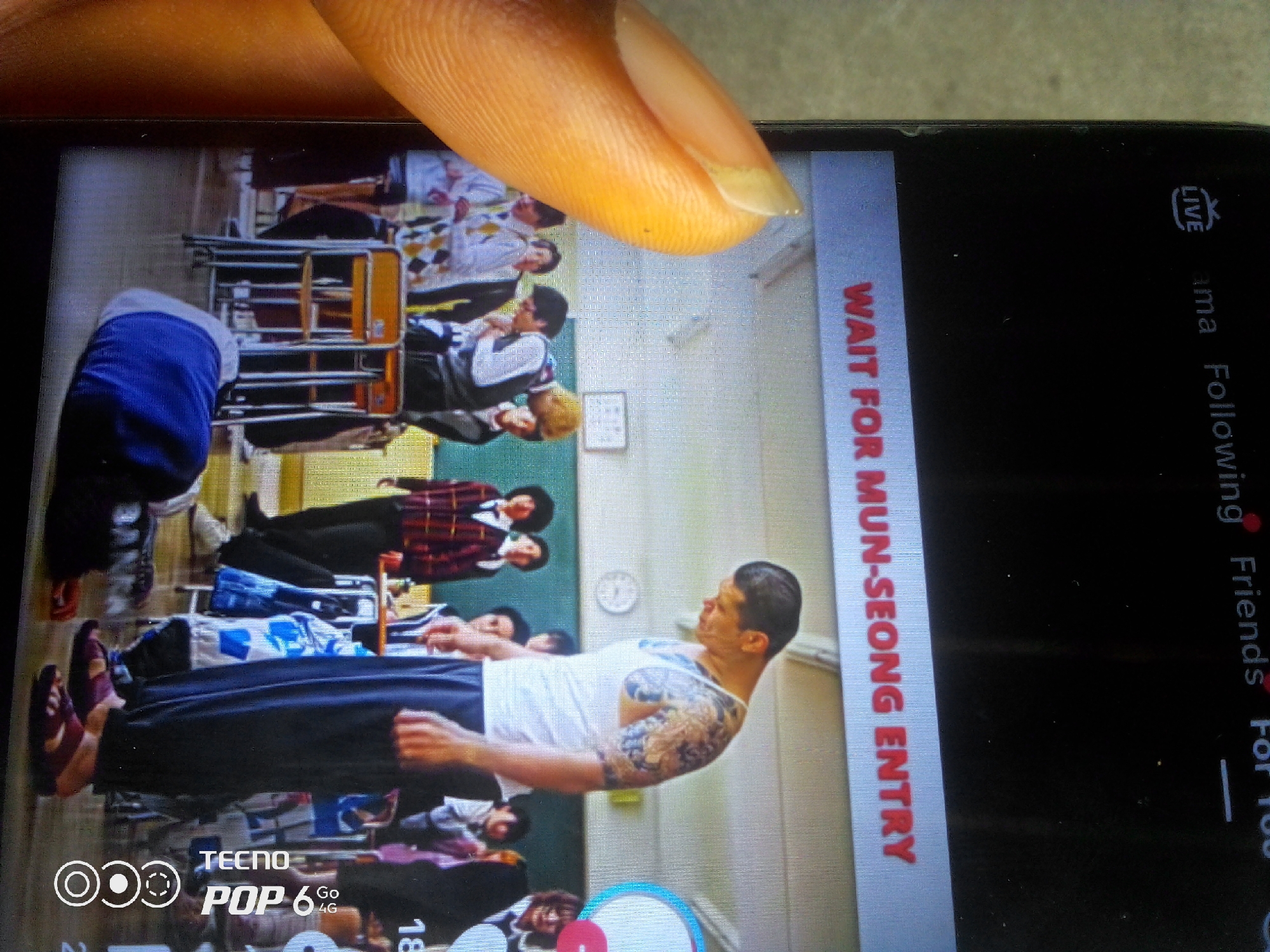 The image captures a screenshot from a phone, displaying a video or live stream. The primary subjects are a group of people, seemingly in a classroom or auditorium setting, as suggested by rows of chairs. One person, a man with tattoos on his arm and chest, is prominently featured in the foreground, appearing to be in motion or striking a pose. He is wearing a white tank top and dark pants. Behind him, several other individuals are visible, some in uniform-like attire, others in casual clothing.
A prominent text overlay on the right side of the screen reads "WAIT FOR MUN-SEONG ENTRY" in red lettering against a blue background. The "LIVE" indicator and other text fragments like "ama Following Friends For 100" suggest a social media or streaming platform interface. At the bottom of the screen, "TECNO POP 6 Go 4G" is visible, identifying the phone model.
A finger is also visible in the top portion of the image, suggesting the photo was taken by someone holding the phone. The overall scene appears to be indoors, with neutral-colored walls and lighting that is neither distinctly bright nor dim, so a specific time of day or weather conditions cannot be determined. The expressions of the people are not clearly discernible due to the resolution and angle. The context provided, "Howayi, Nigeria," places the viewing of this media in that region.