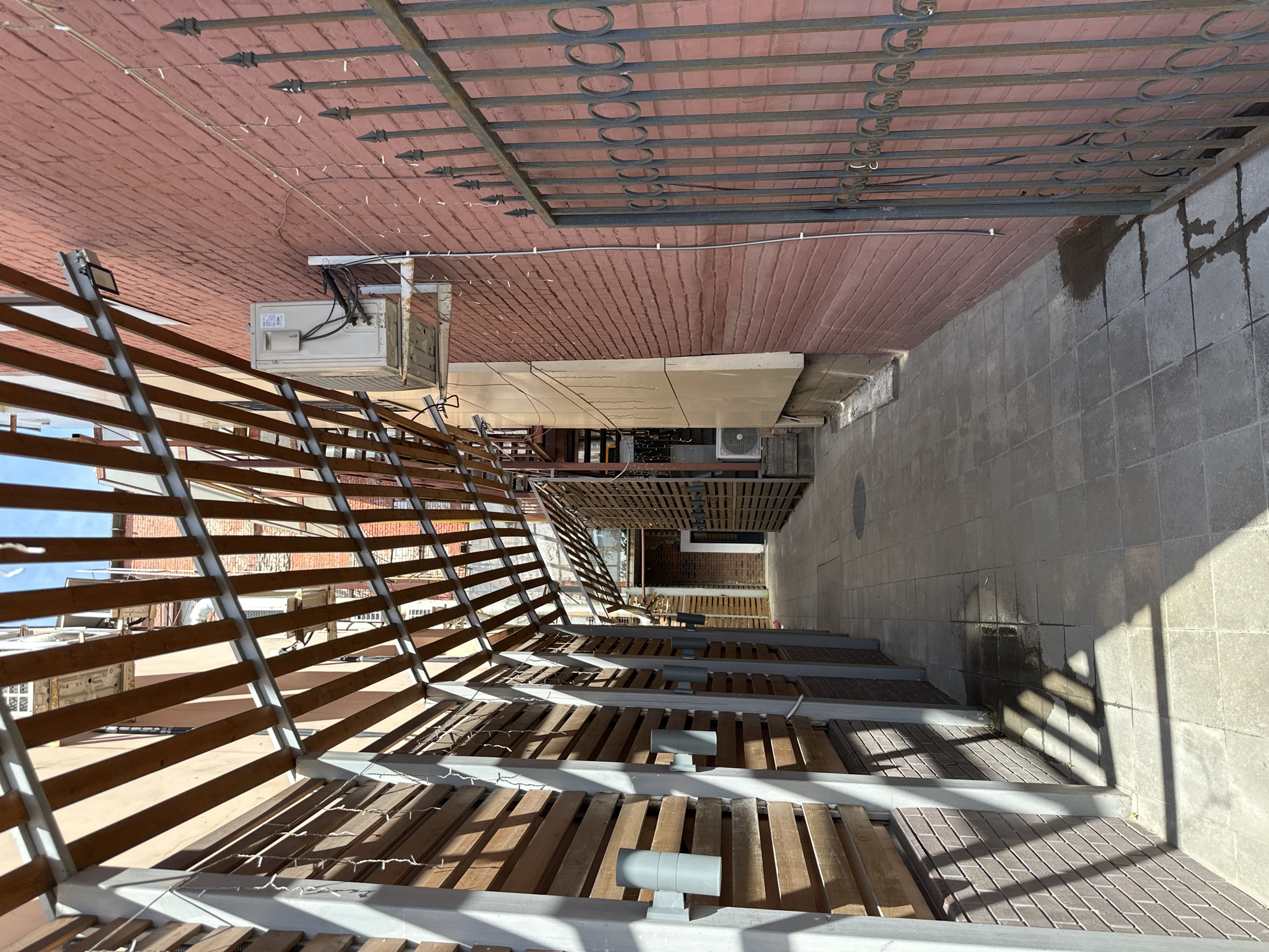 The image shows an outdoor alleyway with a tiled ground surface and brick walls on either side. A wooden slatted overhang with metal supports runs along the alleyway, casting shadows on the ground. Several outdoor light fixtures are attached to the wooden wall on the left. On the right, a rusted metal fence with decorative circular elements runs along the brick wall. An outdoor air conditioning unit is mounted on the brick wall. The scene appears to be in an urban environment, possibly in a commercial or residential area. There are no people visible in the image. The sky is visible at the top of the frame, suggesting it is daytime.