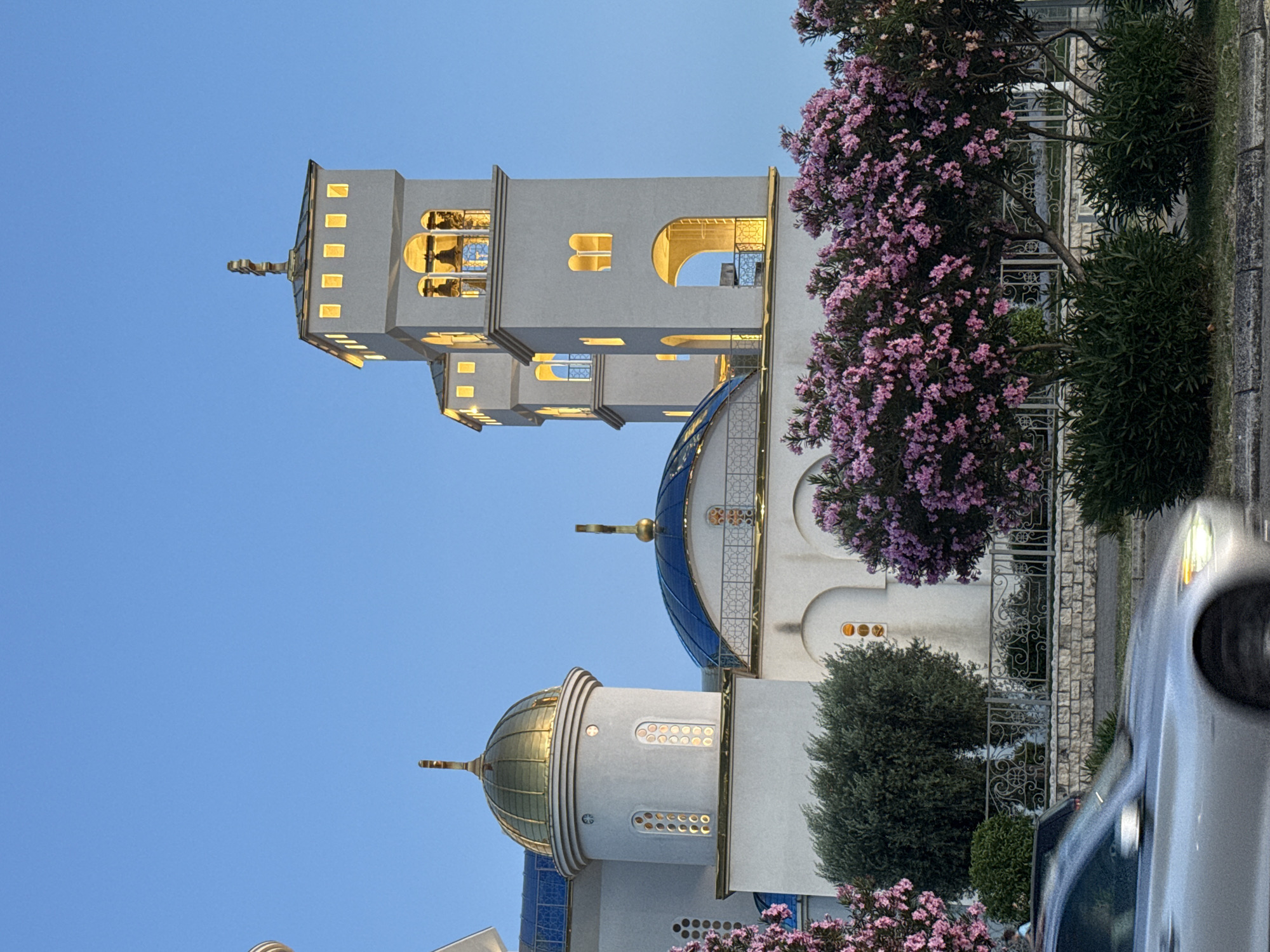 The image shows the Church of Saint Jovan Vladimir in Bar, Montenegro, at dusk. The church is a prominent landmark, characterized by its white facade, golden domes, and a tall bell tower. The sky is a clear, deep blue, indicating the twilight hour. In the foreground, a silver car is blurred as it drives past on a road, suggesting movement. Lush green bushes and trees with vibrant pink flowers flank the church. The architecture features arched windows and decorative elements, with the interior lights of the church glowing warmly through the windows. The bell tower has several levels with open arches, revealing bells within. A cross tops the tallest spire. The scene is serene, showcasing a modern religious structure against a natural and urban backdrop.
