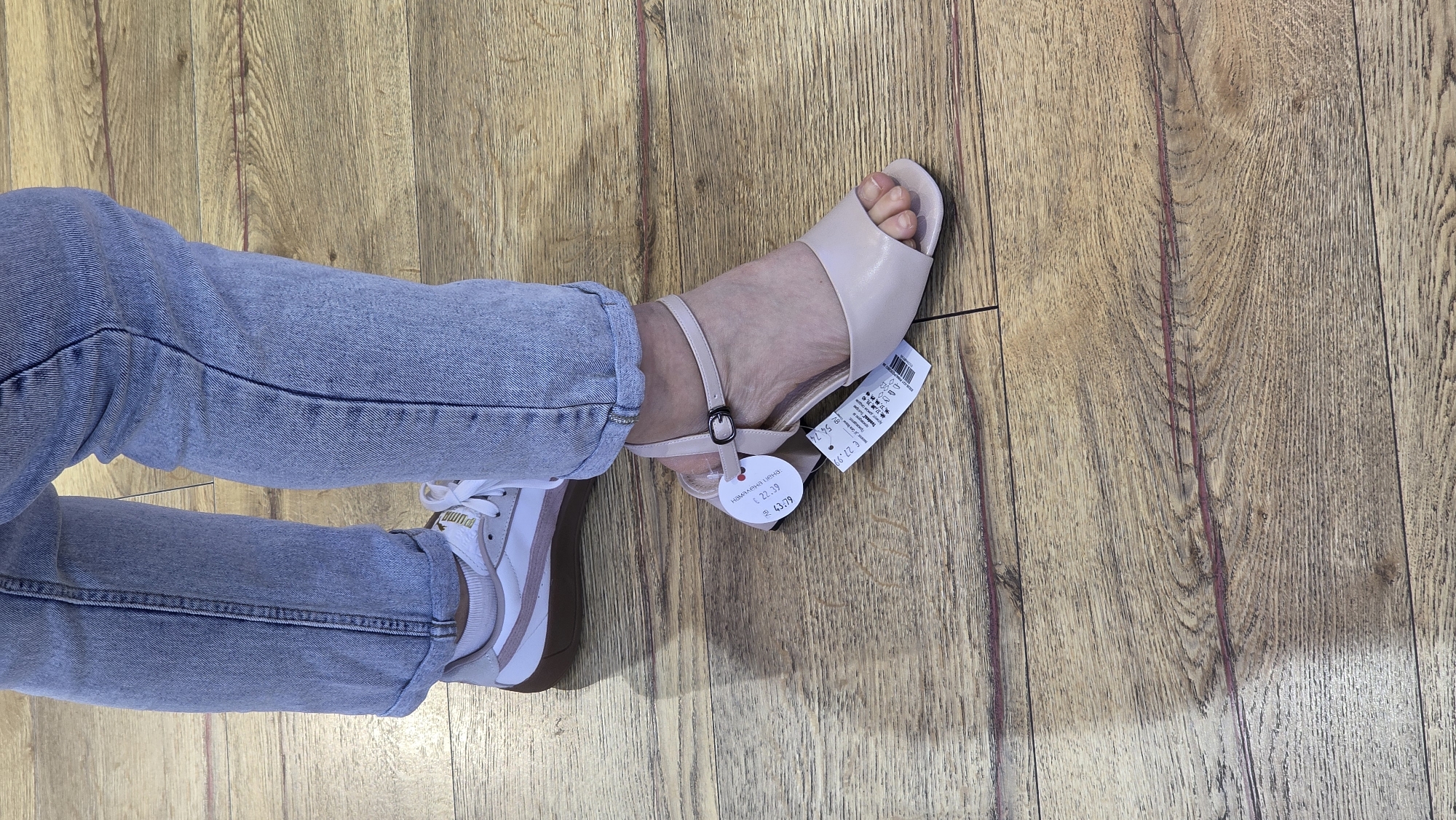 The image depicts a person seated with their legs extended on a wooden floor. The legs are clad in light-wash jeans. One foot is wearing a white and pink Puma sneaker, while the other is adorned with a beige high-heeled sandal that still has its price tags attached. The setting appears to be indoors. Based on the price tag, the location is likely Blagoevgrad, Bulgaria.