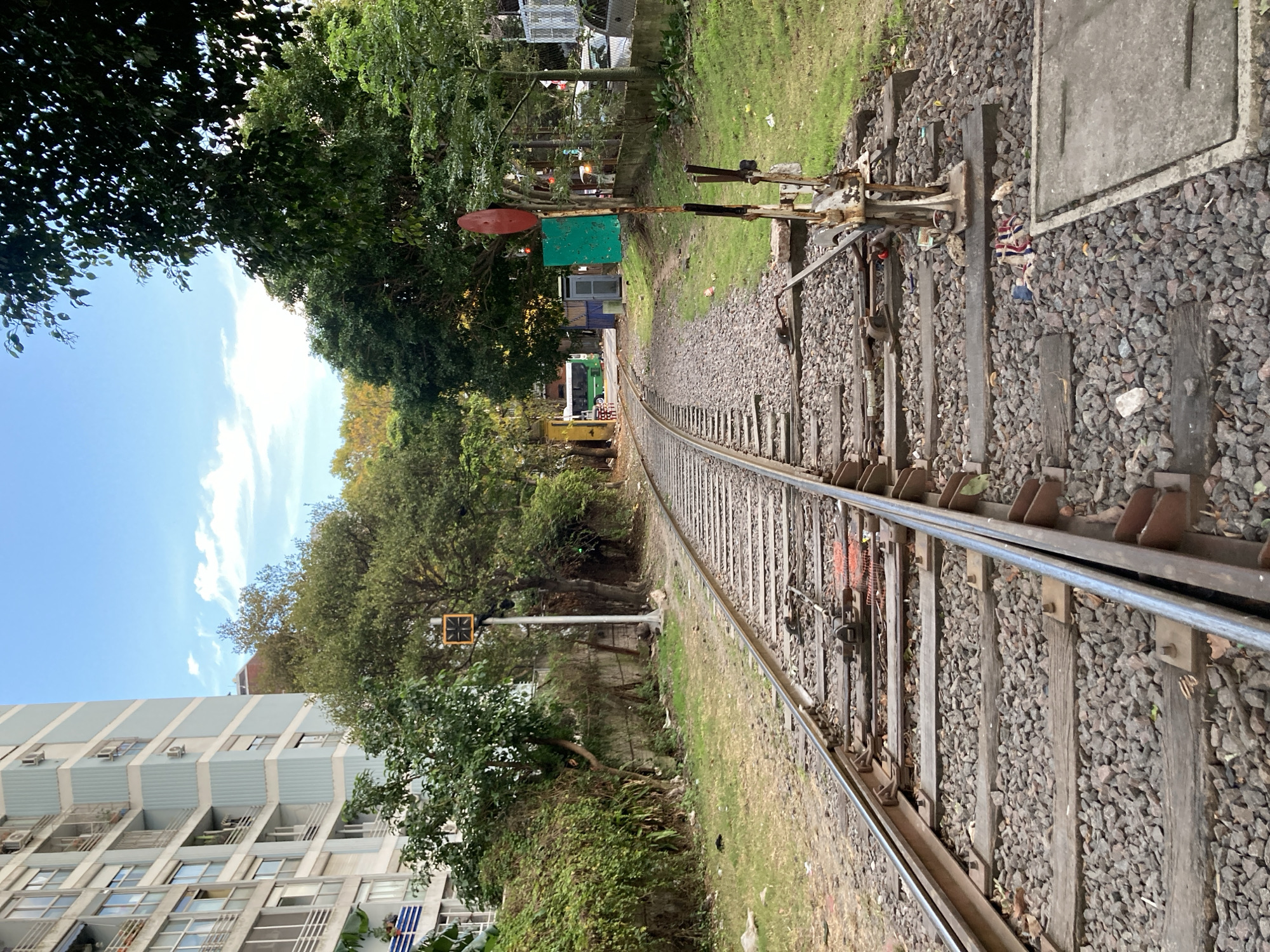 A view of railway tracks curving away from the camera perspective, with a switch mechanism in the foreground. The tracks are surrounded by ballast gravel and vegetation, with a building visible to the lower left. In the distance, there are glimpses of what appears to be a vehicle and other infrastructure. The location is likely within the city of Buenos Aires, Argentina.