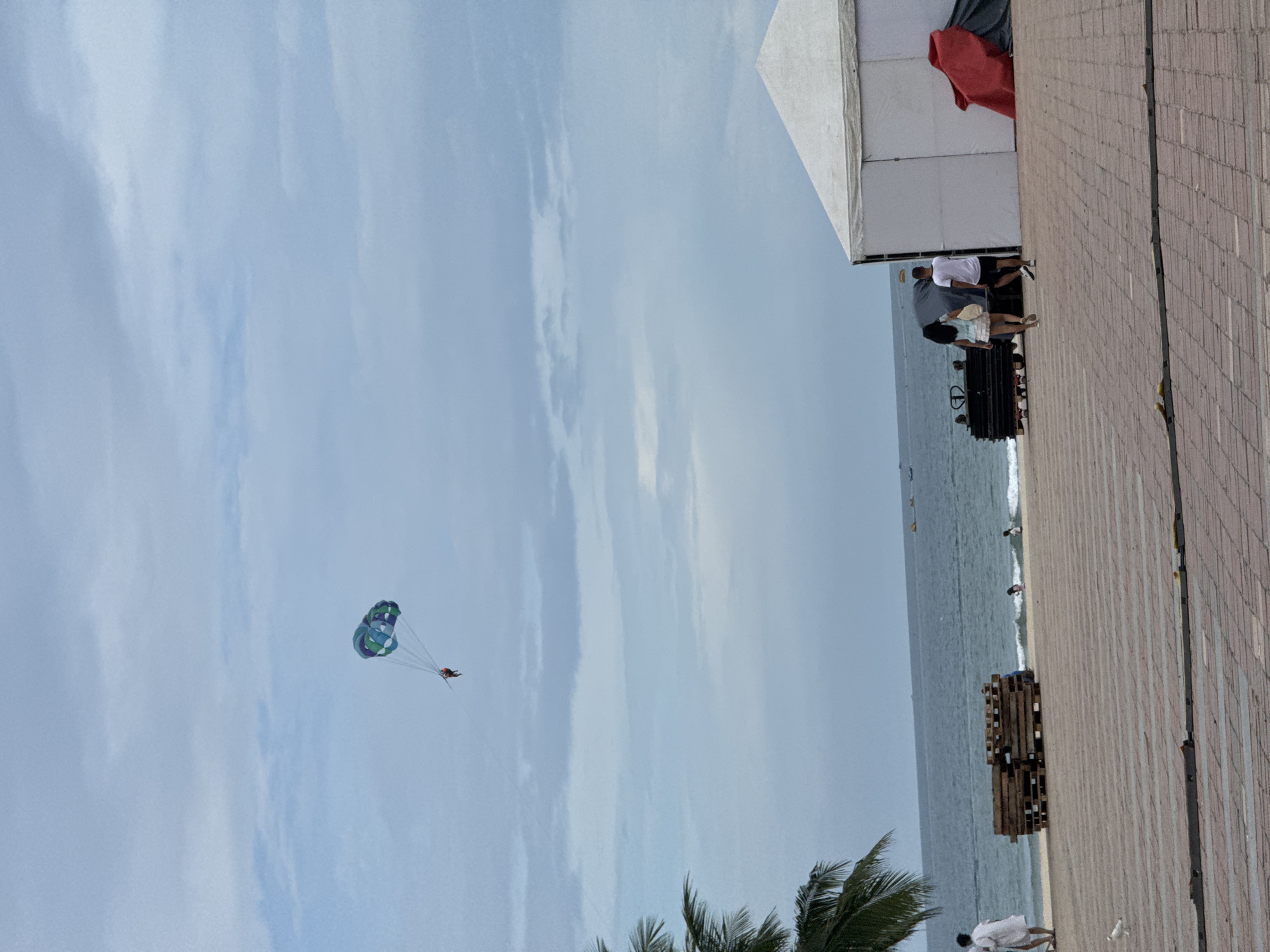 The image captures a beach scene at an unknown location within the city of Đà Nẵng, Vietnam. The sky is overcast with a light gray hue, and scattered clouds are visible. In the distance, a person is parasailing high above the ocean, tethered to a boat that is not fully visible. The ocean itself is a deep blue, with gentle waves lapping at the shore.
On the beach, there are several people. Two individuals, a woman and a man, are walking away from the camera towards the right side of the frame. The woman is wearing a light blue top and a white skirt, carrying a straw bag. The man is in a white t-shirt and black shorts. To their left, there is a large white tent with a triangular roof, partially covering some equipment. A bright red tarp is draped over some dark material near the tent.
Closer to the water, there are stacks of wooden pallets. Further down the beach, two more people, seemingly children, are near the water's edge. A person dressed in a white robe is walking on the beach on the far left of the image. A seagull is visible on the paved walkway in the foreground. The walkway is paved with red and gray rectangular tiles, arranged in a patterned manner. In the background, several small boats are visible on the water. A palm tree frond extends into the frame from the left edge.