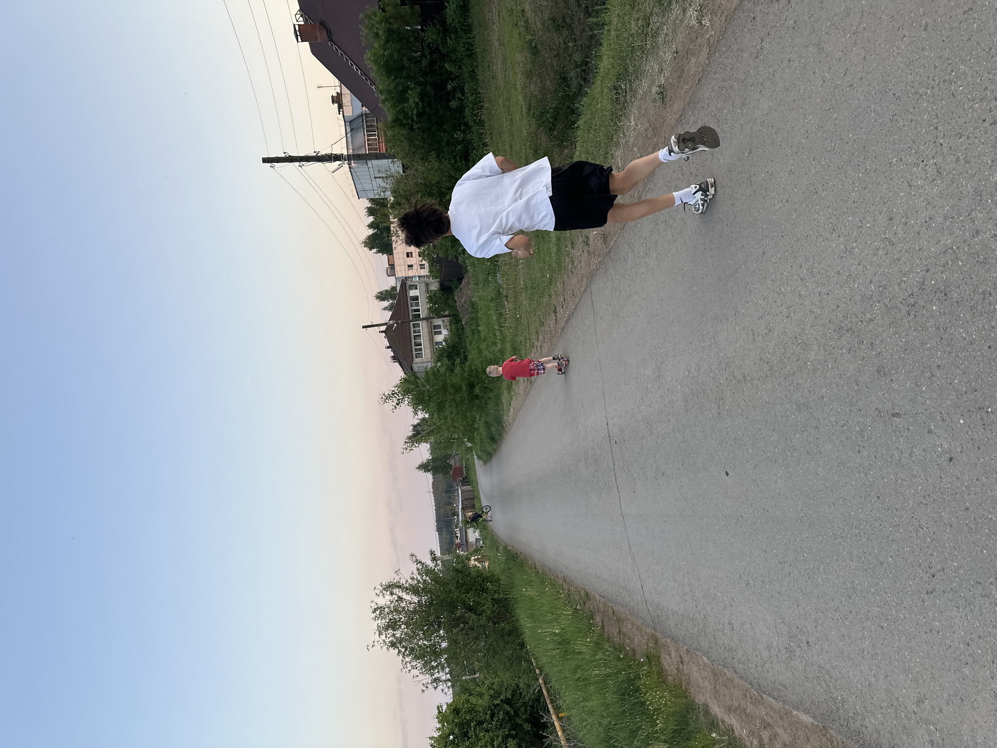 The image shows an asphalt road in Прикамский, Russia. A person in a white shirt and black shorts is running on the road. A younger person in a red shirt is riding a scooter further down the road. There are houses and trees on either side of the road. The sky is light blue and pink, suggesting it is dawn or dusk. A utility pole can be seen near the houses on the left. A bicycle can be seen further down the road on the left.
