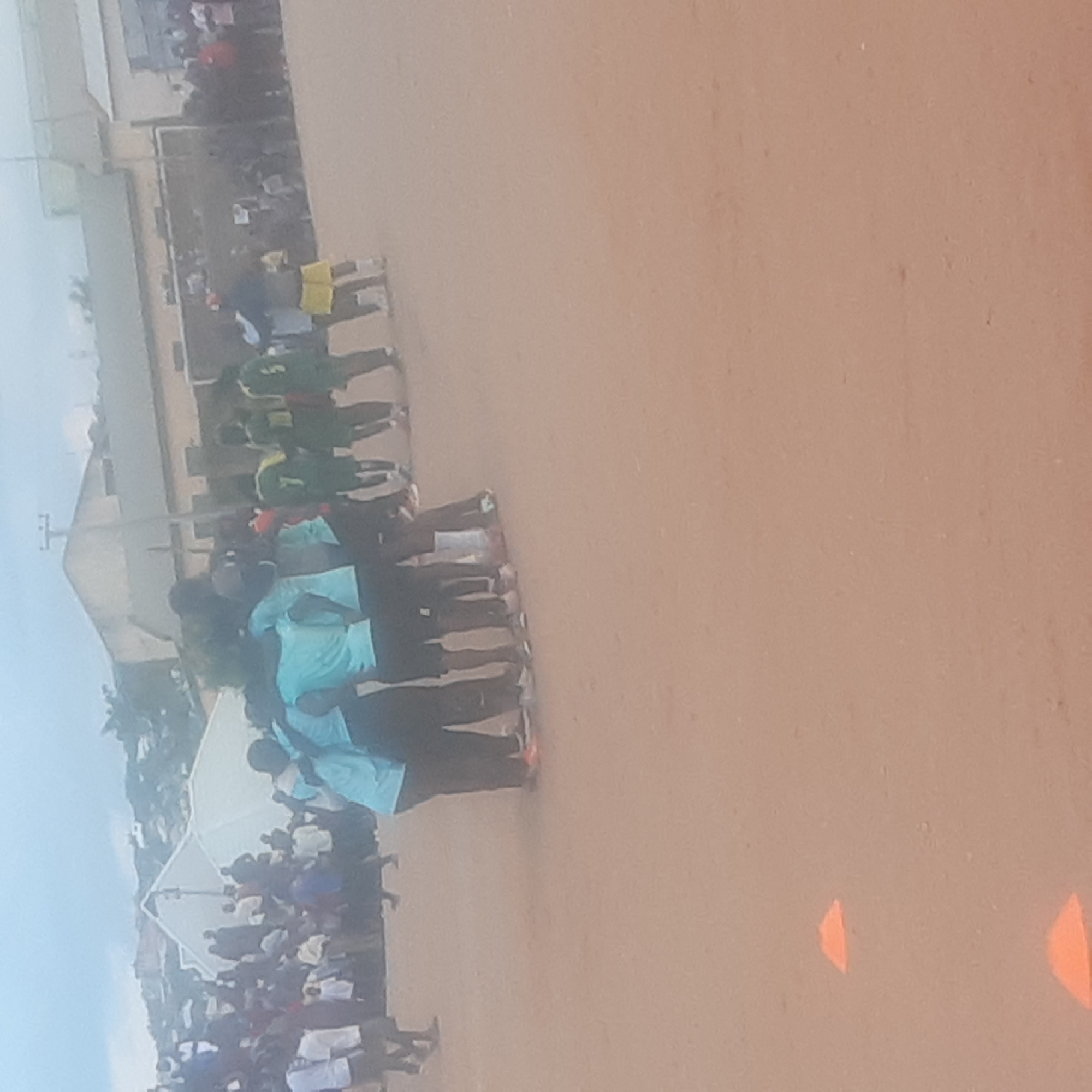 The image depicts a group of young people, likely a sports team, huddled together in a circle on a dirt field. They are wearing athletic attire, with some in light blue shirts and black pants, and others in green shirts and shorts. One individual in the foreground is wearing yellow shorts and a dark blue shirt. In the background, a crowd of spectators is visible, along with some white tents and buildings that suggest an event is taking place. The overall setting appears to be an outdoor sports facility. The image seems to have been taken in Jos, Nigeria, where such events are common.