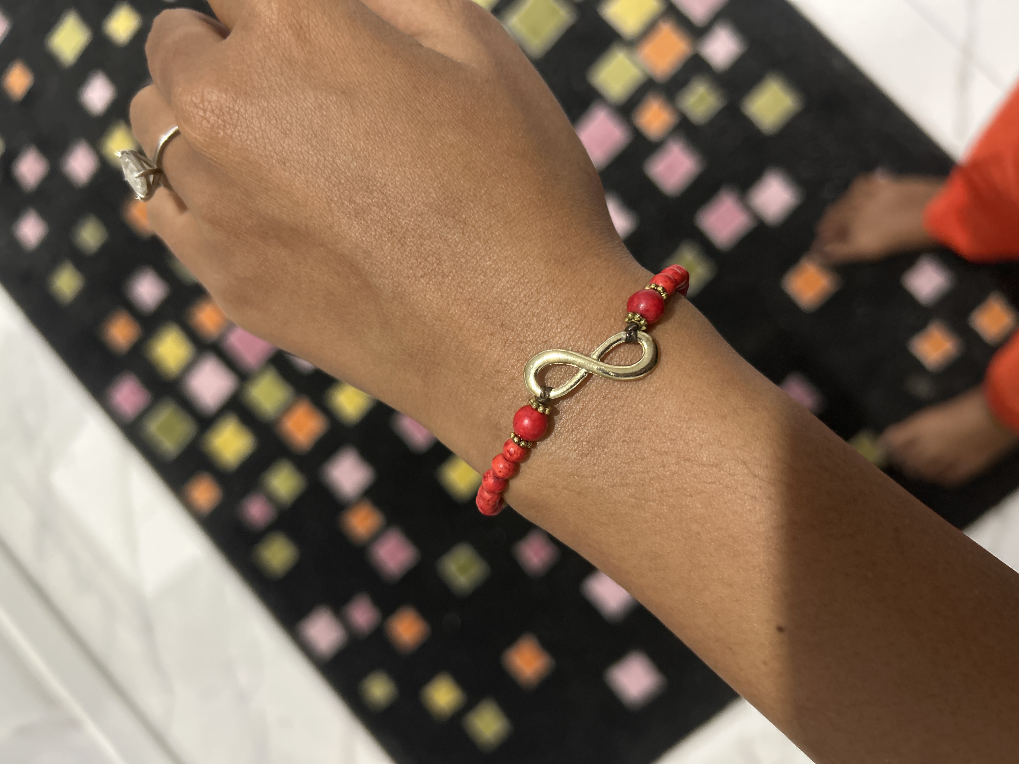 A close-up shot focuses on a person's wrist adorned with a red beaded bracelet featuring a gold infinity symbol charm. The person wears a large, faceted diamond ring on their index finger. The background is blurred, showing a black surface with a pattern of colorful squares in shades of pink, yellow, orange, and green. The floor appears to be tiled with white marble. The scene is indoors, with soft lighting that creates shadows on the skin. The image is framed from the perspective of looking down at the wrist and hand.