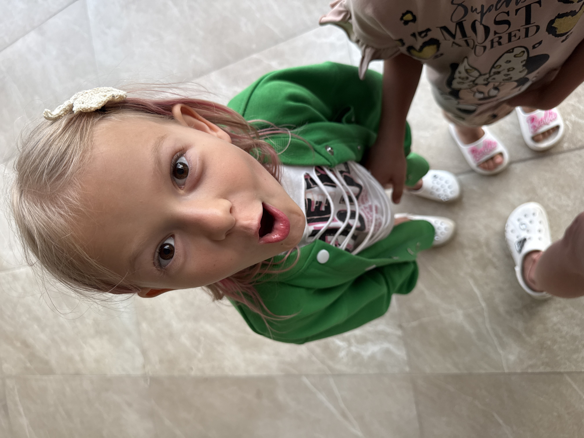A young girl with blonde hair and pink streaks, wearing a green jacket over a white shirt, looks up at the camera with her mouth open in surprise. She has a small white bow clipped to her hair. She is standing on a light-colored, marble-like floor. Another person's legs and feet, wearing a pink shirt with Mickey Mouse print and white sandals, are visible to the right of the girl, reaching out towards her. Another pair of white clogs are visible on the floor. The perspective is from a high angle, looking down at the girl.