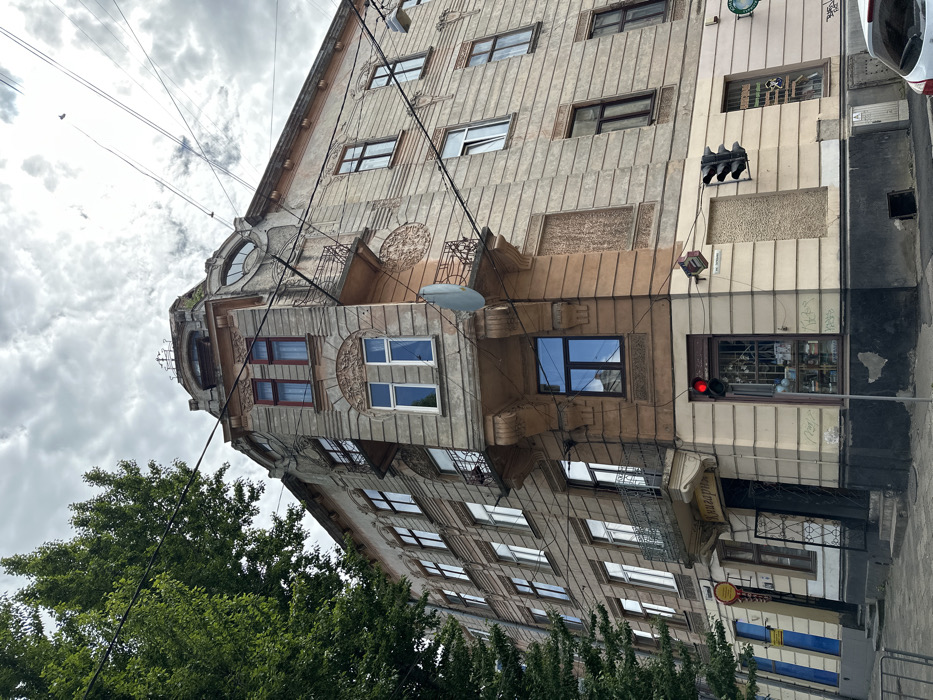 The image shows a corner building in Solonka, Ukraine, at latitude 49.829 and longitude 24.032. The building is a multi-story structure with a large storefront on the corner. The building has a distinctive architectural style with ornate details and a curved corner. The storefront is closed with a black metal gate and a sign that reads "Магазин". There are windows and balconies on the building's facade, which is painted in a pale yellow color. The building's exterior shows signs of wear and tear, including chipped paint and cracked plaster. The sidewalk in front of the building is paved with grey concrete and appears to be deserted. The street is lined with trees and a red traffic light is illuminated. A lone blue car is parked in the distance. The sky is overcast with a few wispy clouds. Power lines extend overhead, creating a network of intersecting cables across the image.